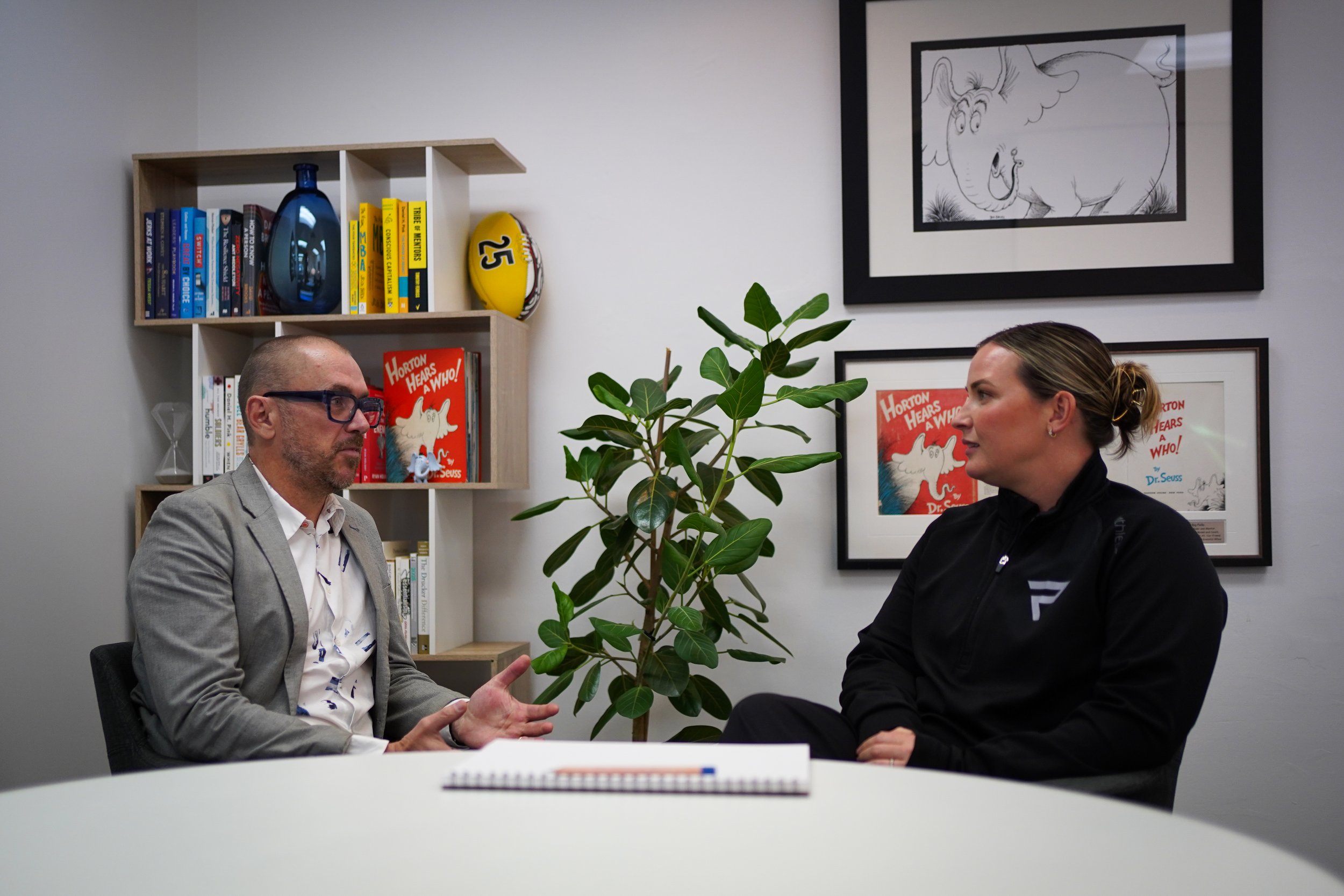 Two people, a man with glasses and a woman with a ponytail, are sitting at a white table in a room. They are having a conversation. Behind them is a bookshelf with books, decorative items, and a yellow football. A large potted plant is in the foreground. Framed artwork and a poster featuring "Horton Hears a Who!" are hanging on the wall.