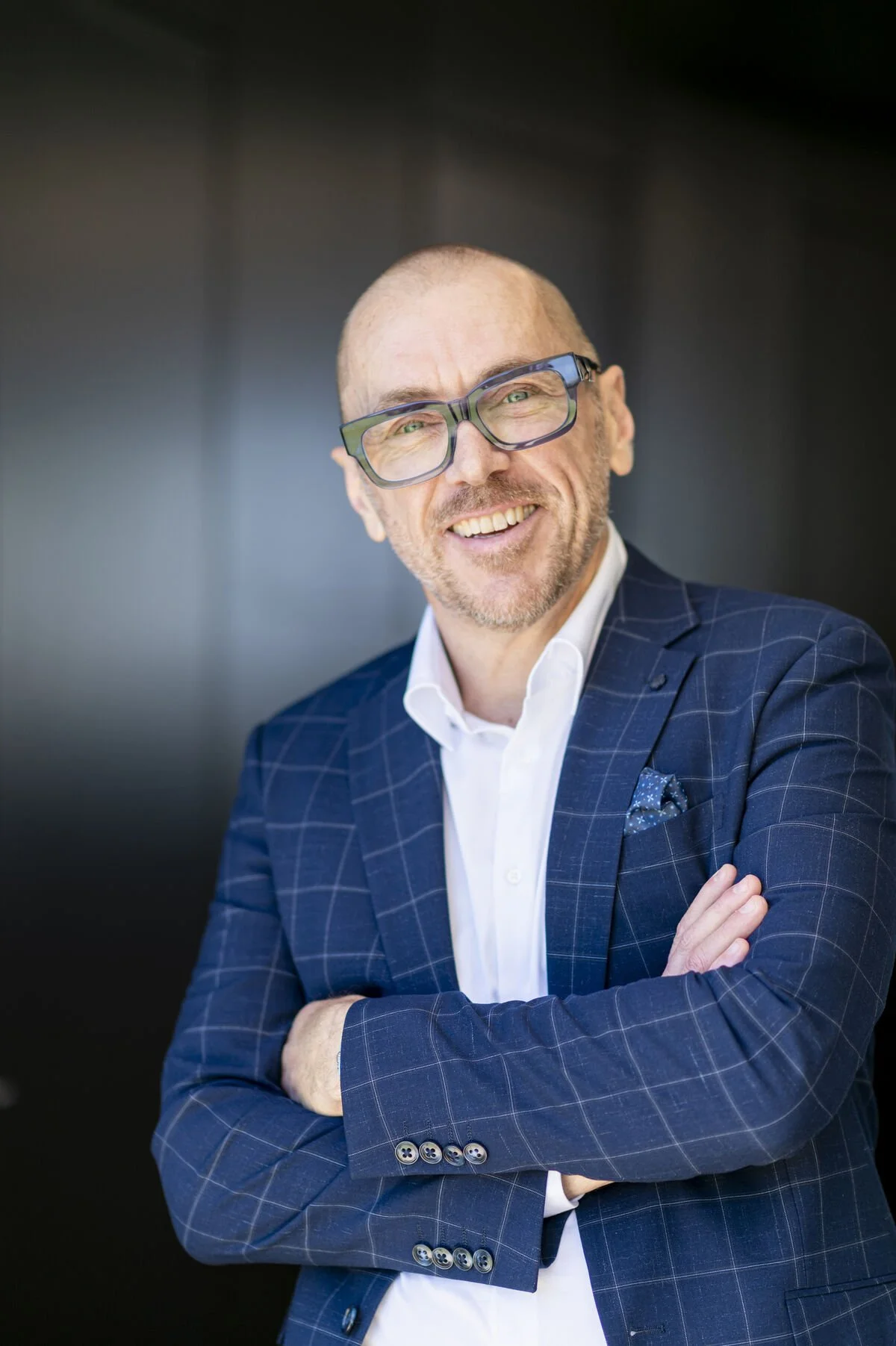 A man with glasses, bald head, and a beard, wearing a navy blue checkered suit and white shirt, smiling with arms crossed against a dark background.