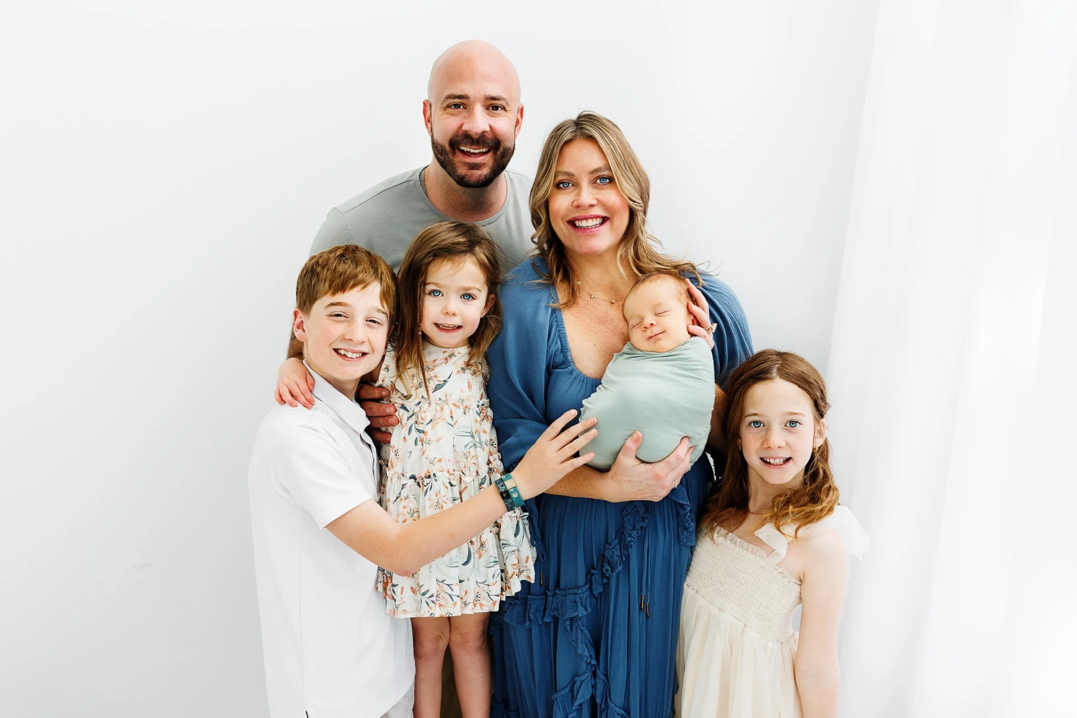 Family of six, including two adults and four children, smiling and gathered together in a well-lit room with a white background. The mother holds a sleeping baby, while the other children stand close by.