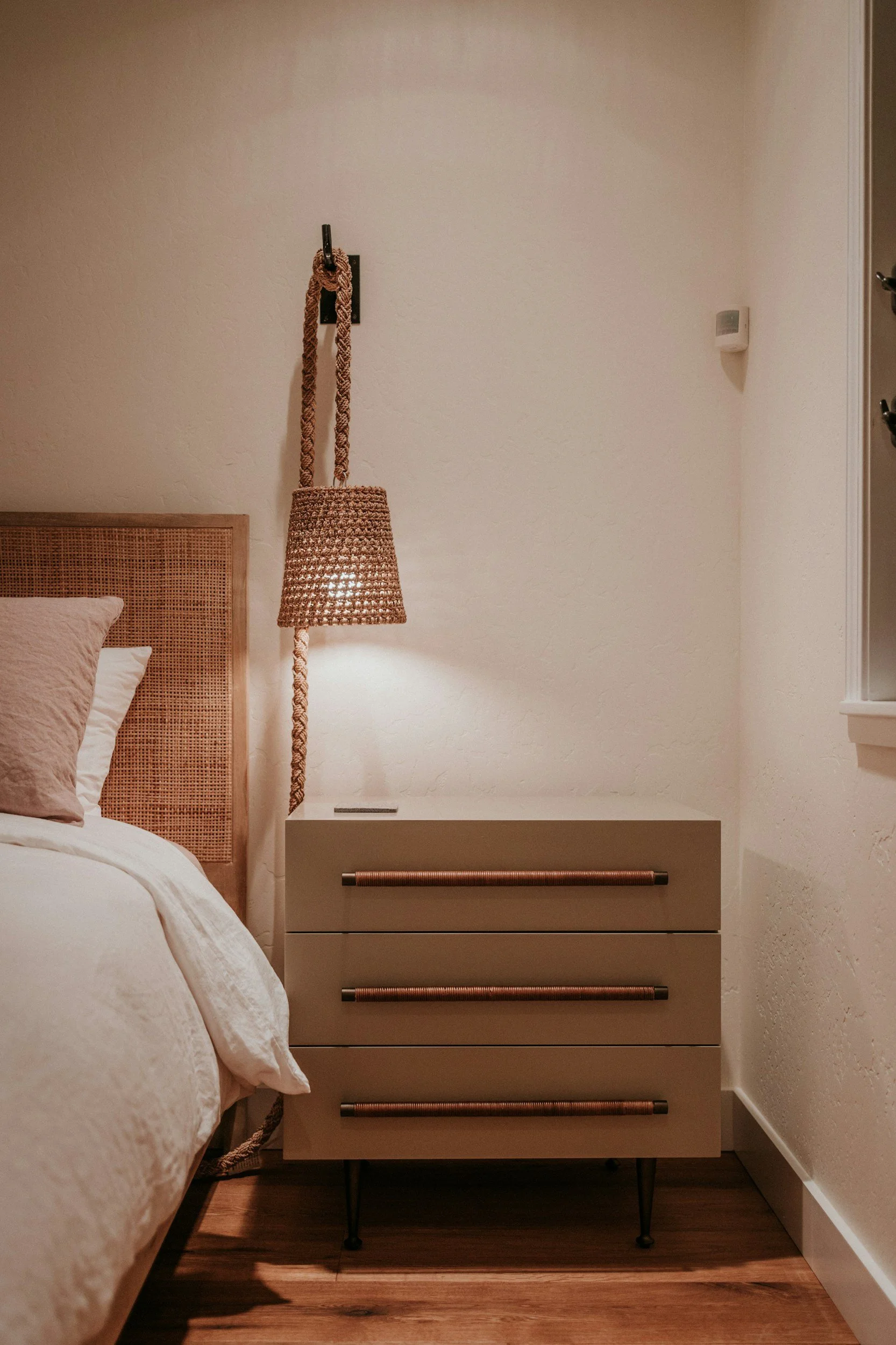 A bedroom with a bed, rattan headboard, beige bedding, a nightstand, and a wicker-style hanging lamp with a braided cord