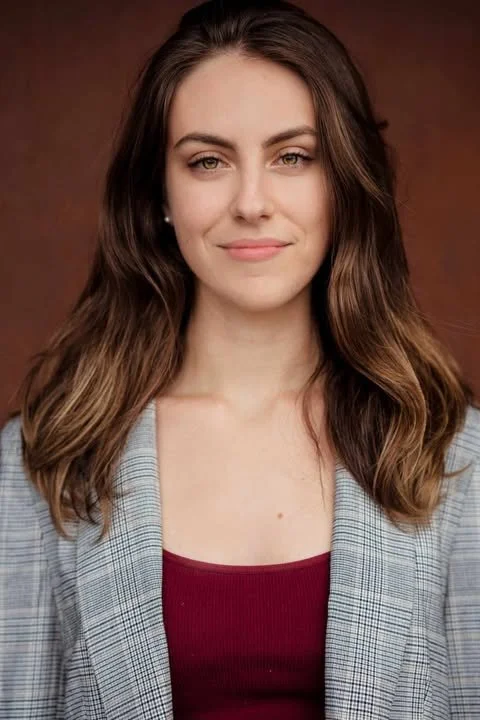 The face behind MJ Vine Marketing, Melissa, who has with long wavy brown hair and fair skin, wearing a gray plaid blazer over a maroon top, smiling slightly with a neutral expression against a brown background.