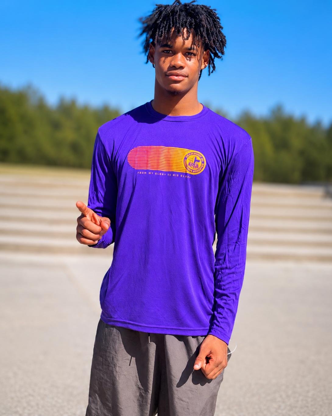 Young man with dreadlocks wearing a purple sports shirt with a logo and text, standing outdoors on a sunny day.