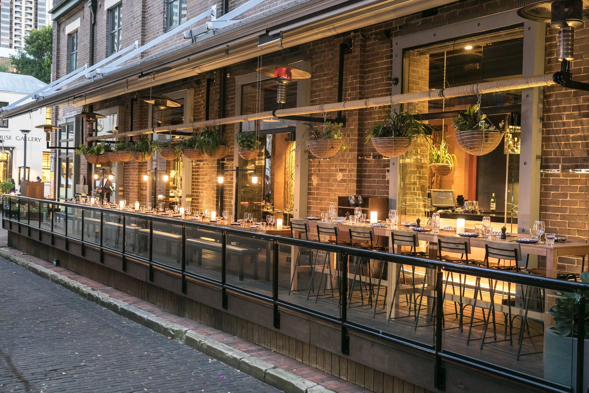 Exterior view of a restaurant with large glass windows, a brick facade, and hanging potted plants. Indoor dining setup with tables, chairs, and candle-lit dinner arrangements visible through the windows.