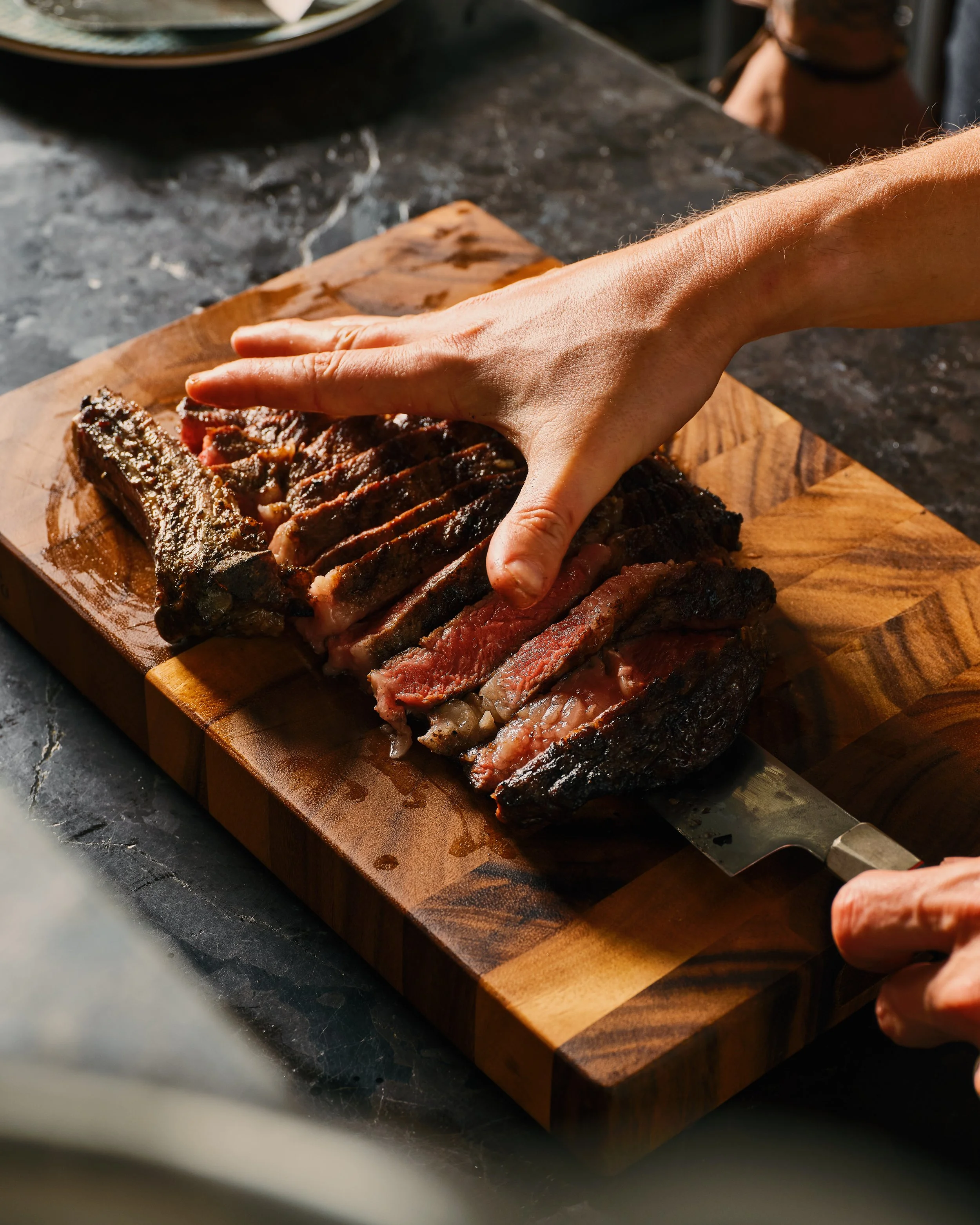 A person slicing cooked meat on a wooden cutting board with a large knife, on a dark countertop.