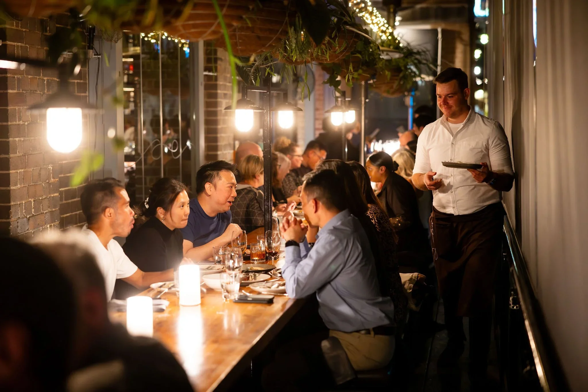 A group of people dining at a restaurant with warm lighting, arranged along a long table. A waiter in a white shirt is serving them, standing beside a curtain. The restaurant has exposed brick walls, hanging plants, and string lights.