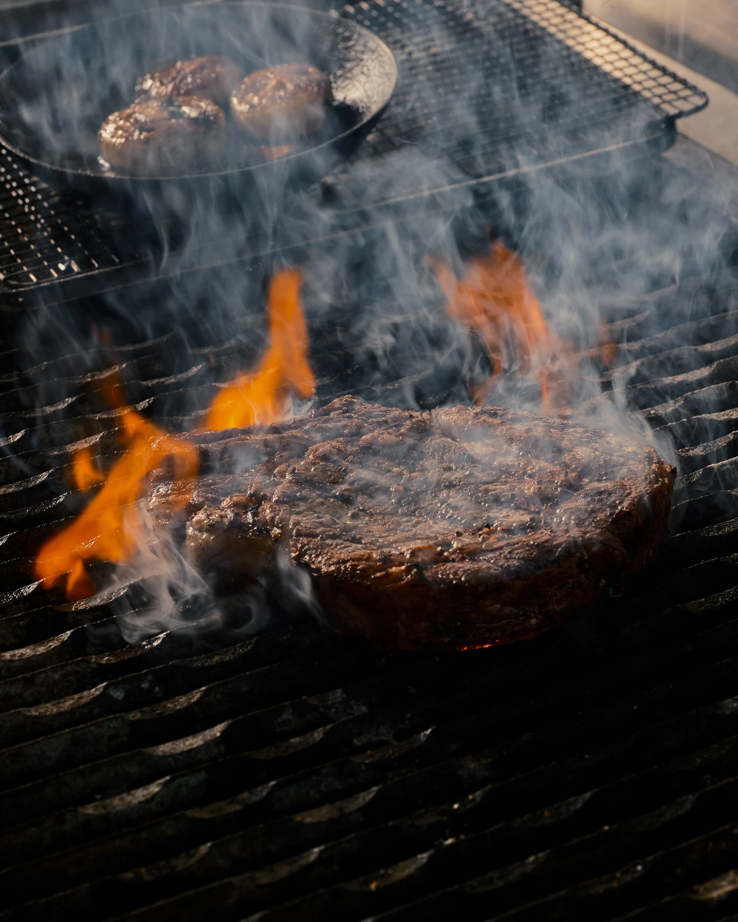 A grill with burning flames, a cast iron skillet with meatballs, and steam rising from the skillet.