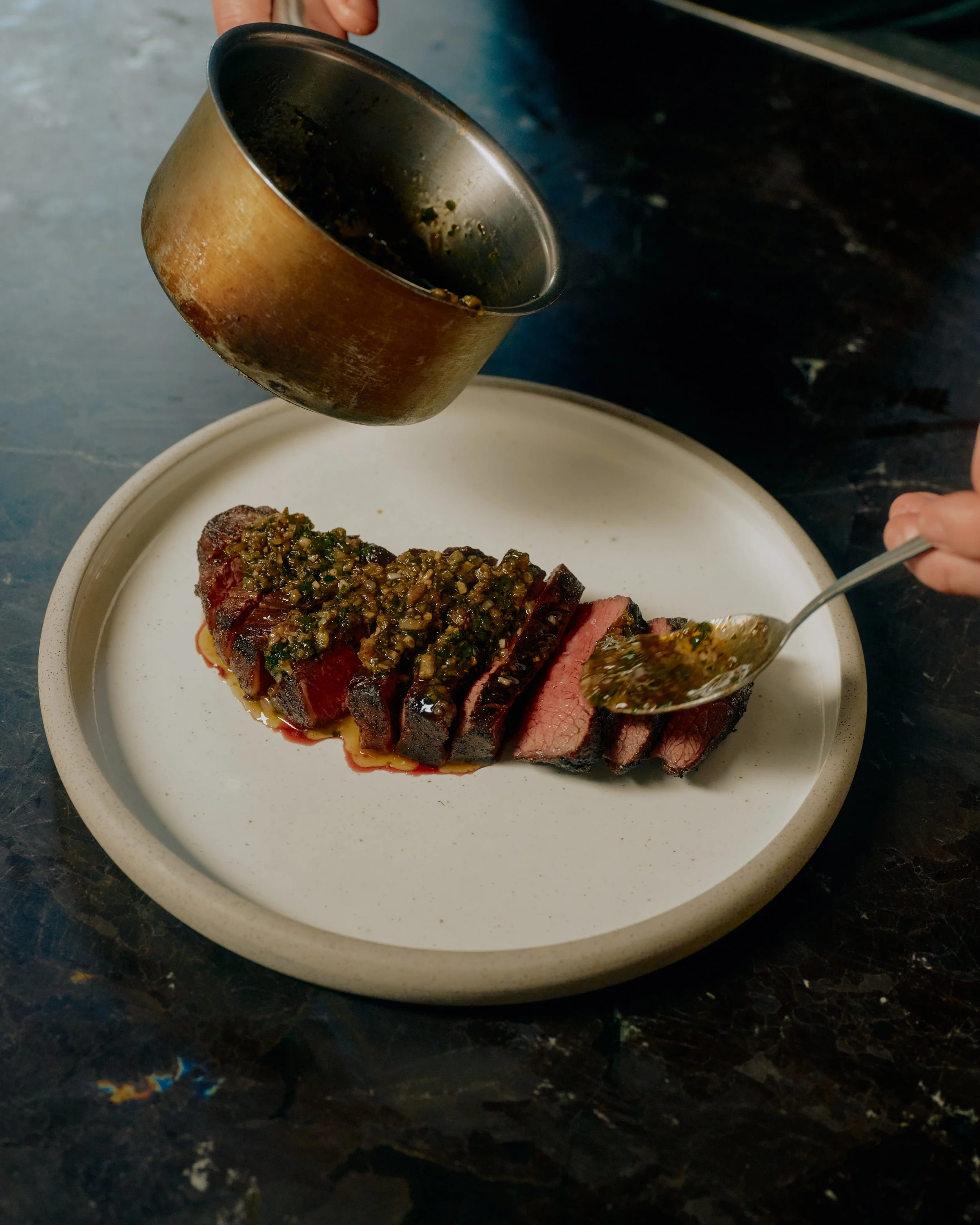 A person pouring a dark sauce over a sliced medium-rare steak topped with chimichurri on a white plate.