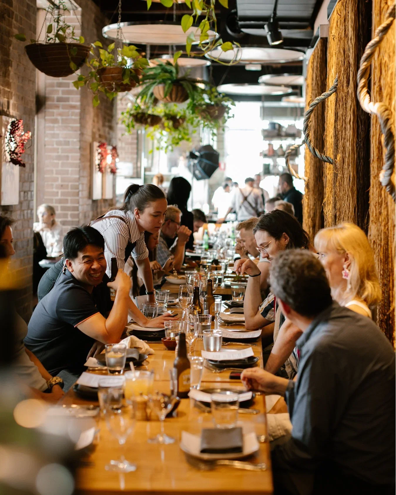 People dining at a long table in a restaurant with hanging plants and brick walls.