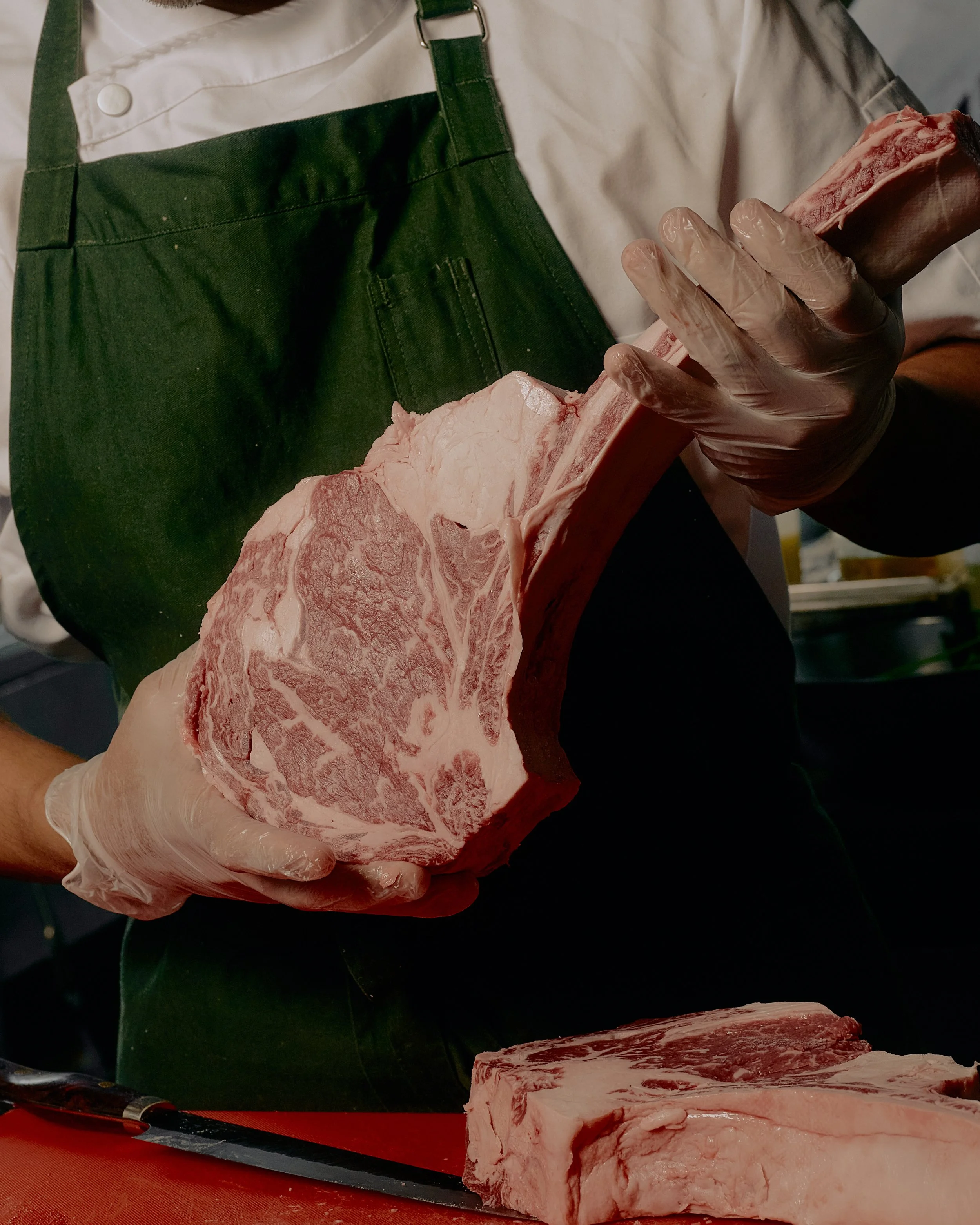 Person wearing gloves and a green apron cutting raw beef cuts on a cutting board with a chef's knife.