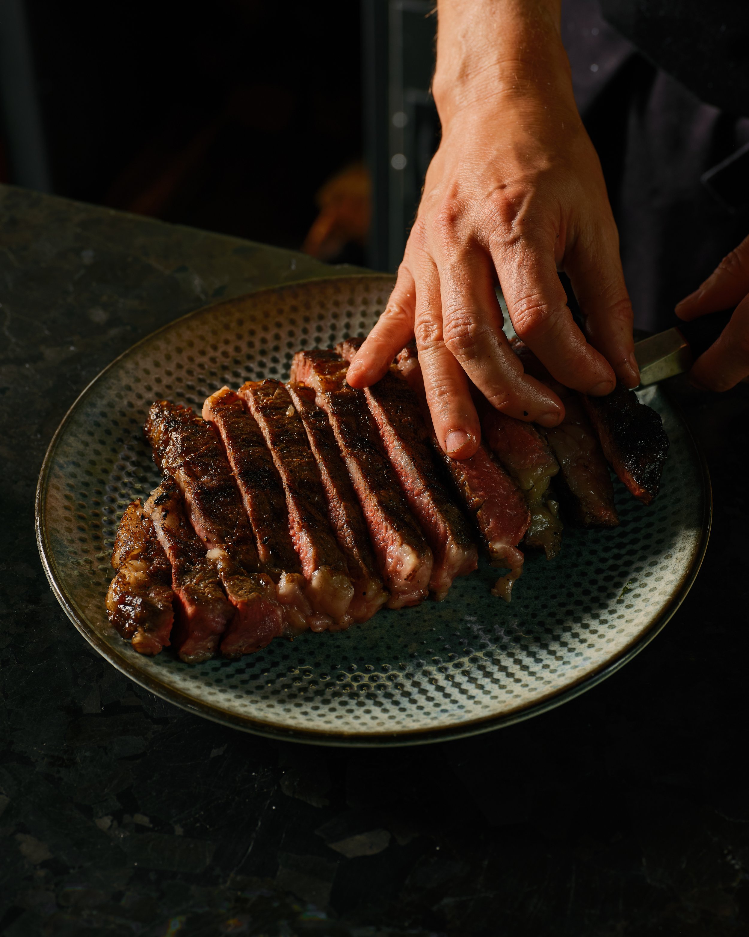 A person slicing cooked medium-rare steak on a dark green plate, with a dark background.