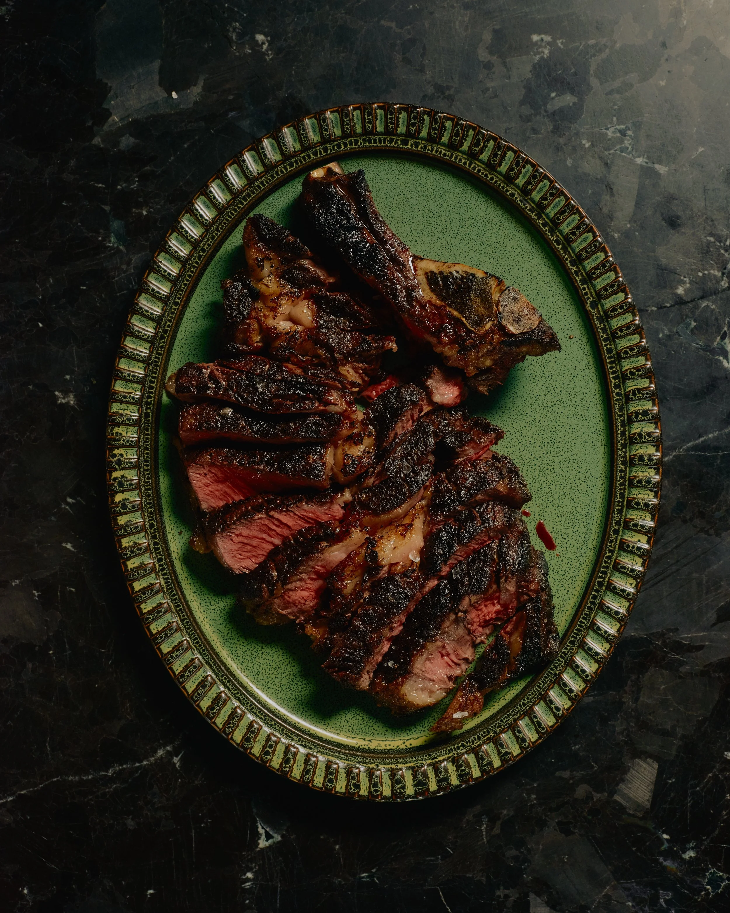 Cooked steak on a green and gold decorative oval plate on a black marble surface.