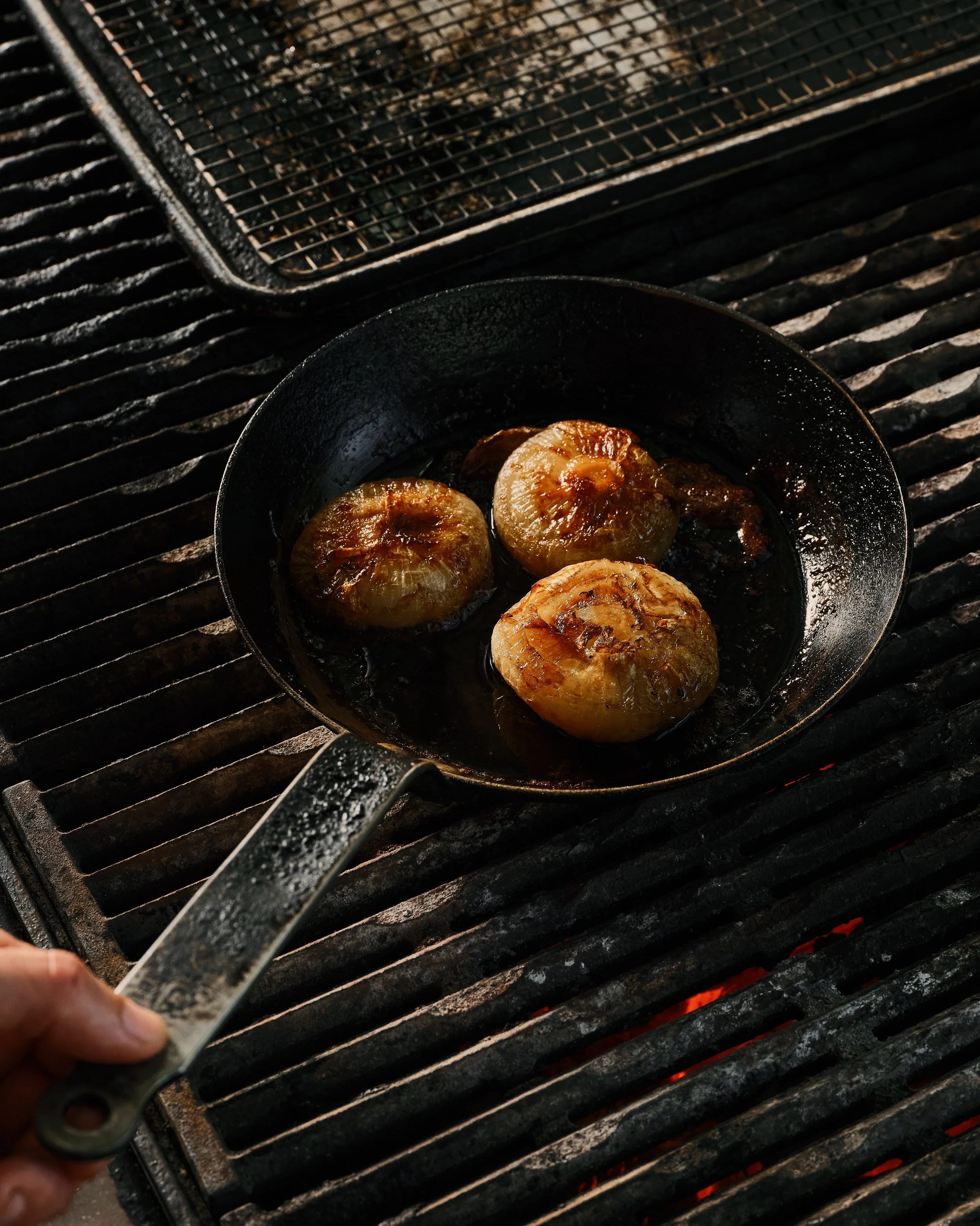 Three scallops being cooked in a black cast-iron skillet over a grill. Part of a grill grate is visible in the background.