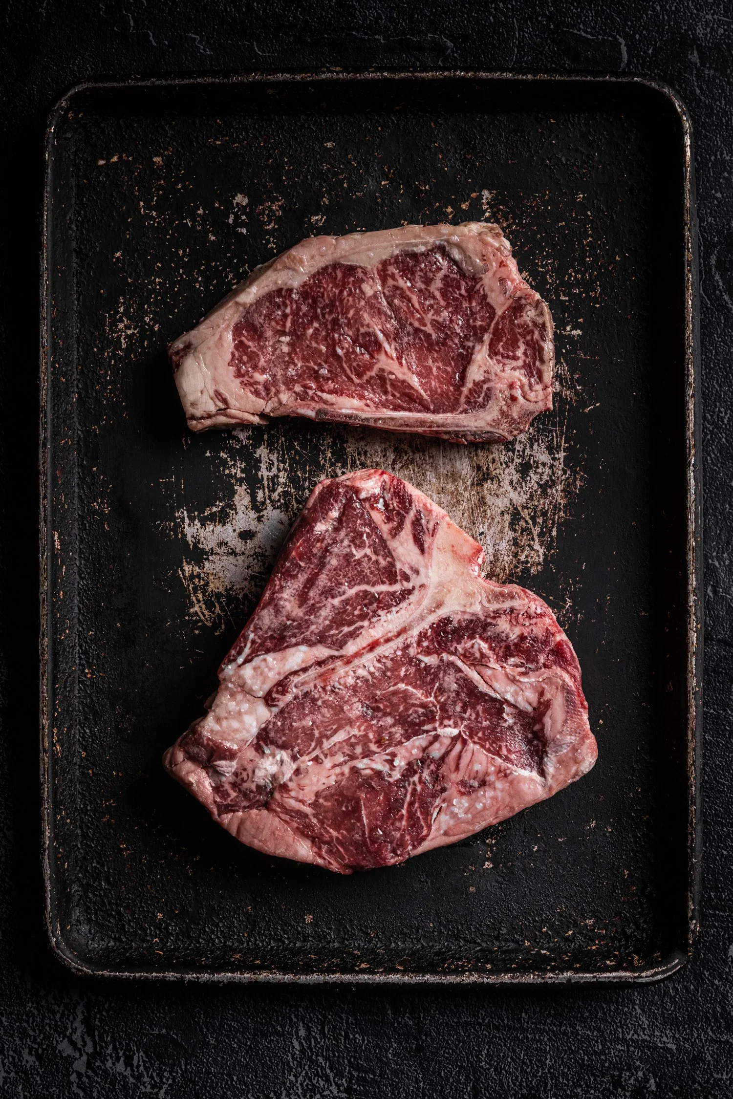 Three raw beef steaks on a black baking sheet, seasoned with salt and spices.