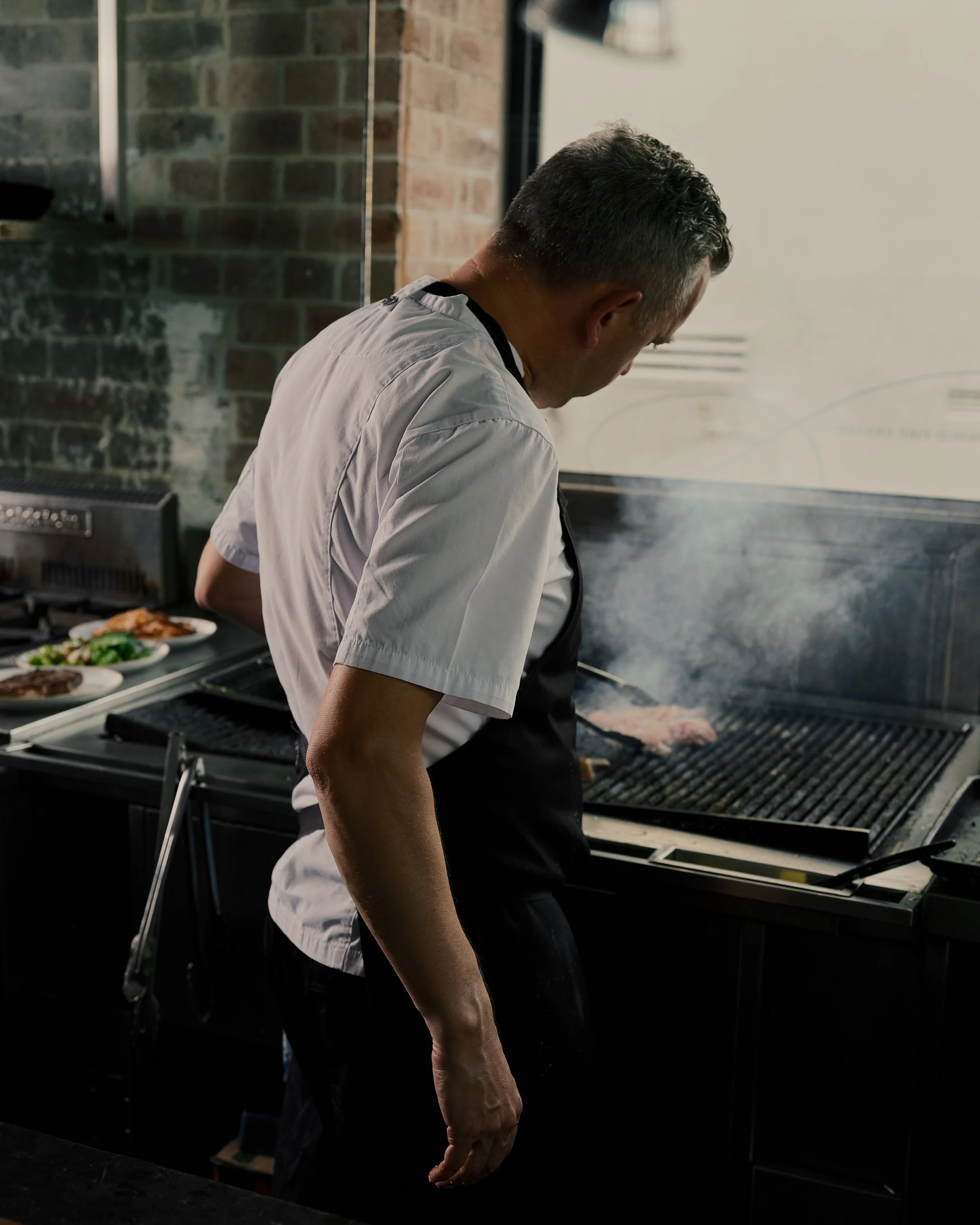 A man in a white shirt and black apron is grilling meat on a barbecue grill in a kitchen or restaurant setting, with plates of cooked food on a counter behind him.