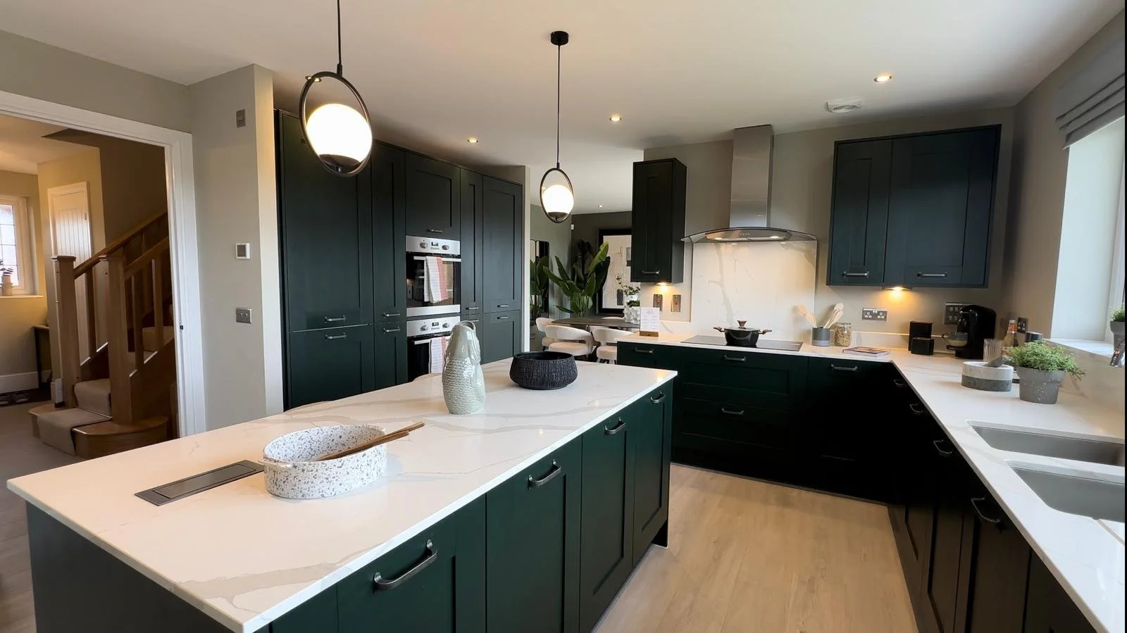 Modern kitchen with dark green cabinets, white countertops, and the island in the center. There are pendant lights above the island, and a dining area with white chairs in the background. Kitchen appliances and decorative items are on the counters.