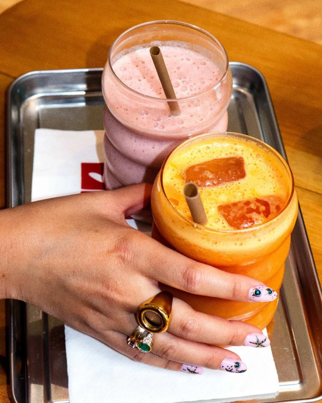 Deux smoothies, un rose et un orange, dans des verres avec pailles, posé sur un plateau en métal avec une main portant l'orange, tous sur une nappe blanche sur une table en bois.
