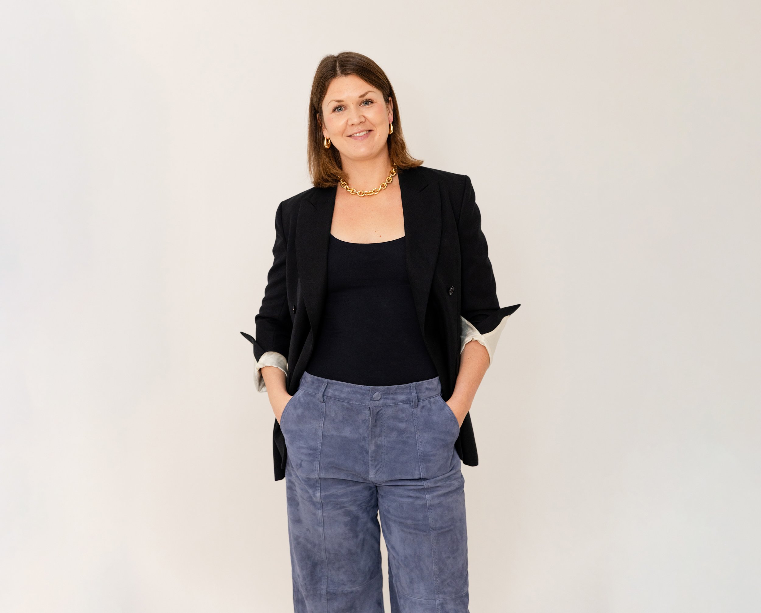 Kvinna i svart kavaj och blå jeans står mot en vit vägg, ler lätt.