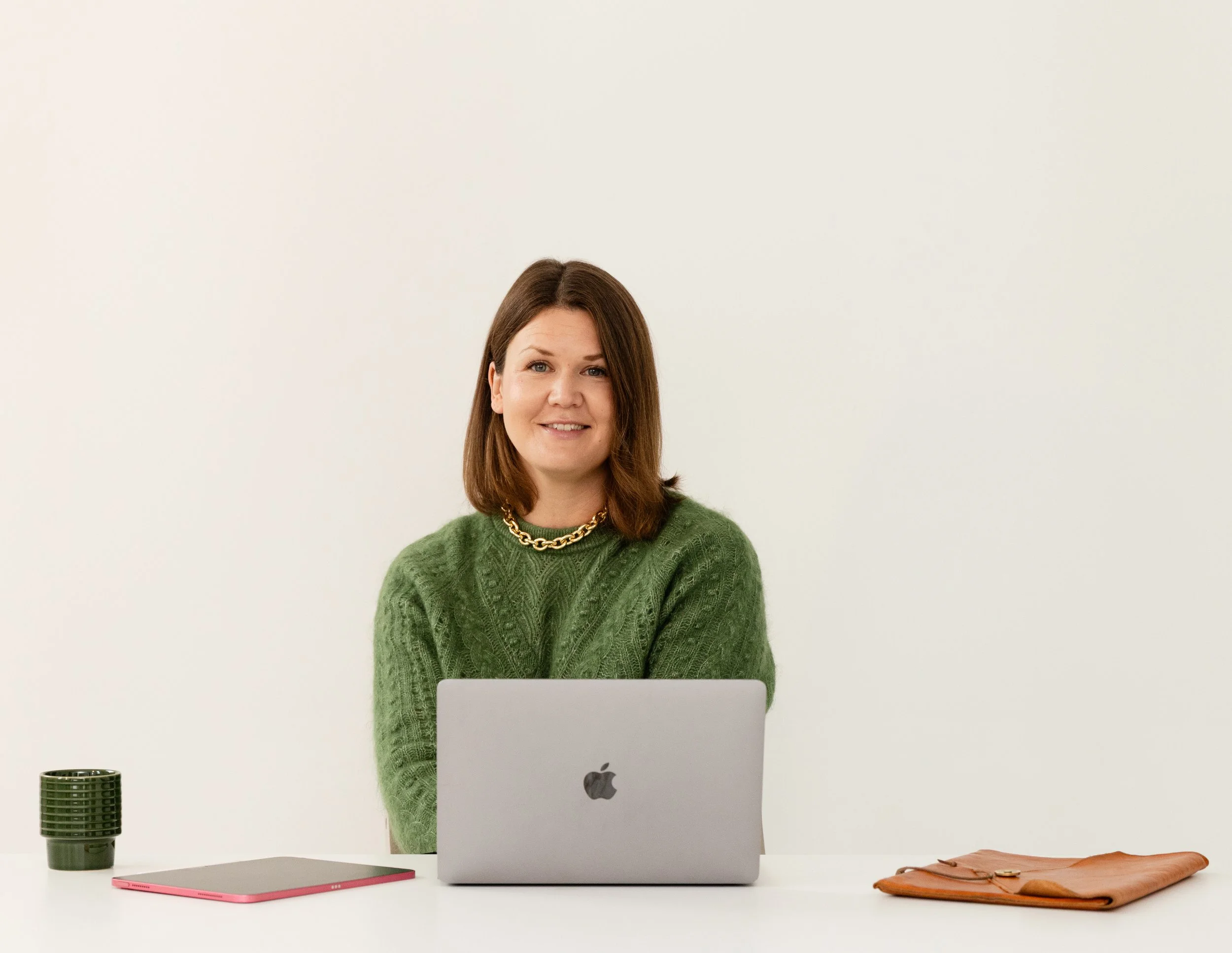 En kvinna med brunt hår, grönt stickat/svampat tröja, guldhalsband, sitter framför en Apple MacBook och tittar mot kameran. På skrivbordet finns också en svart kaffekopp, en rosa anteckningsbok, ett orange plånbok och en svart mikrofon. Bakgrund är vit.