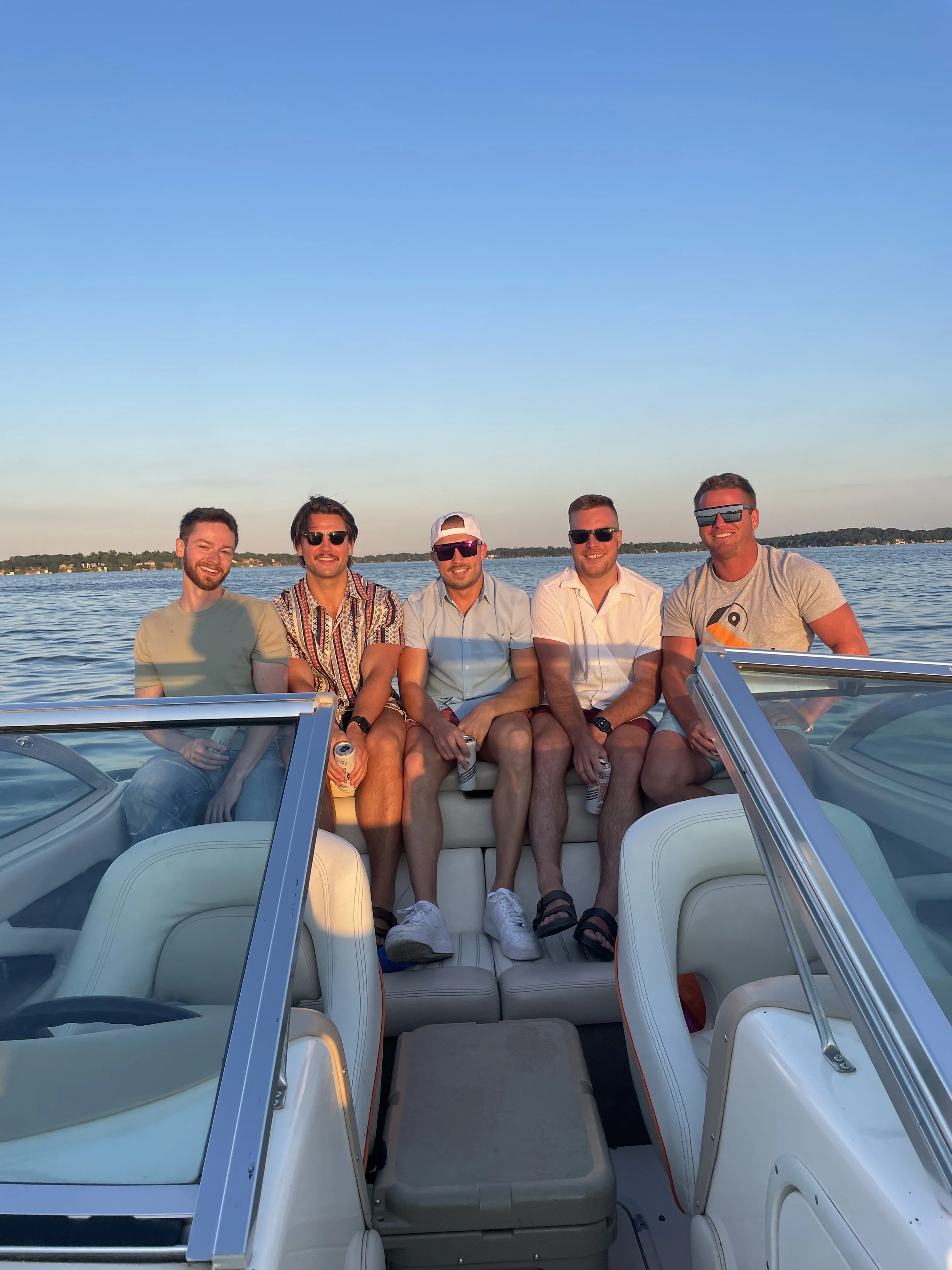 Five young men sitting on a boat with water and trees in the background during sunset, all wearing sunglasses and casual clothes, smiling at the camera.