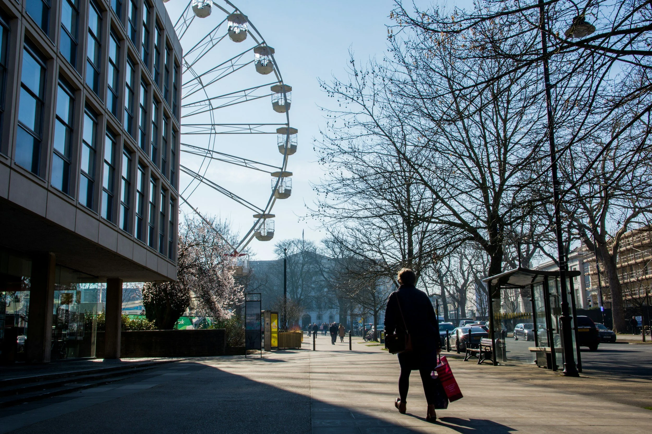 A street scene with a large Ferris wheel, leafless trees, and buildings. A person with a shopping bag is walking on the sidewalk near a bus stop on a sunny day.