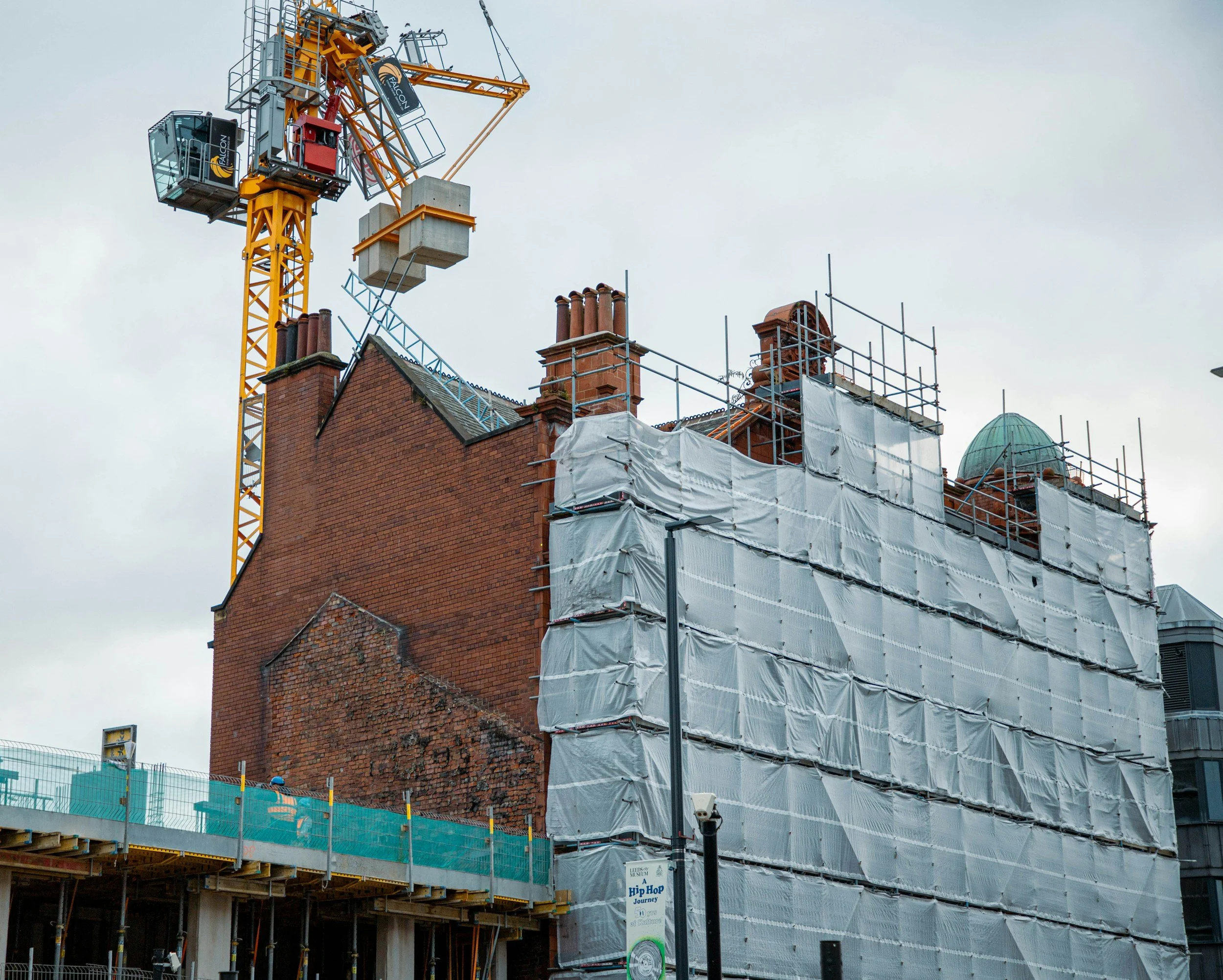 Construction site with a yellow tower crane lifting concrete blocks onto an old brick building, which is covered in scaffolding and white protective sheeting.