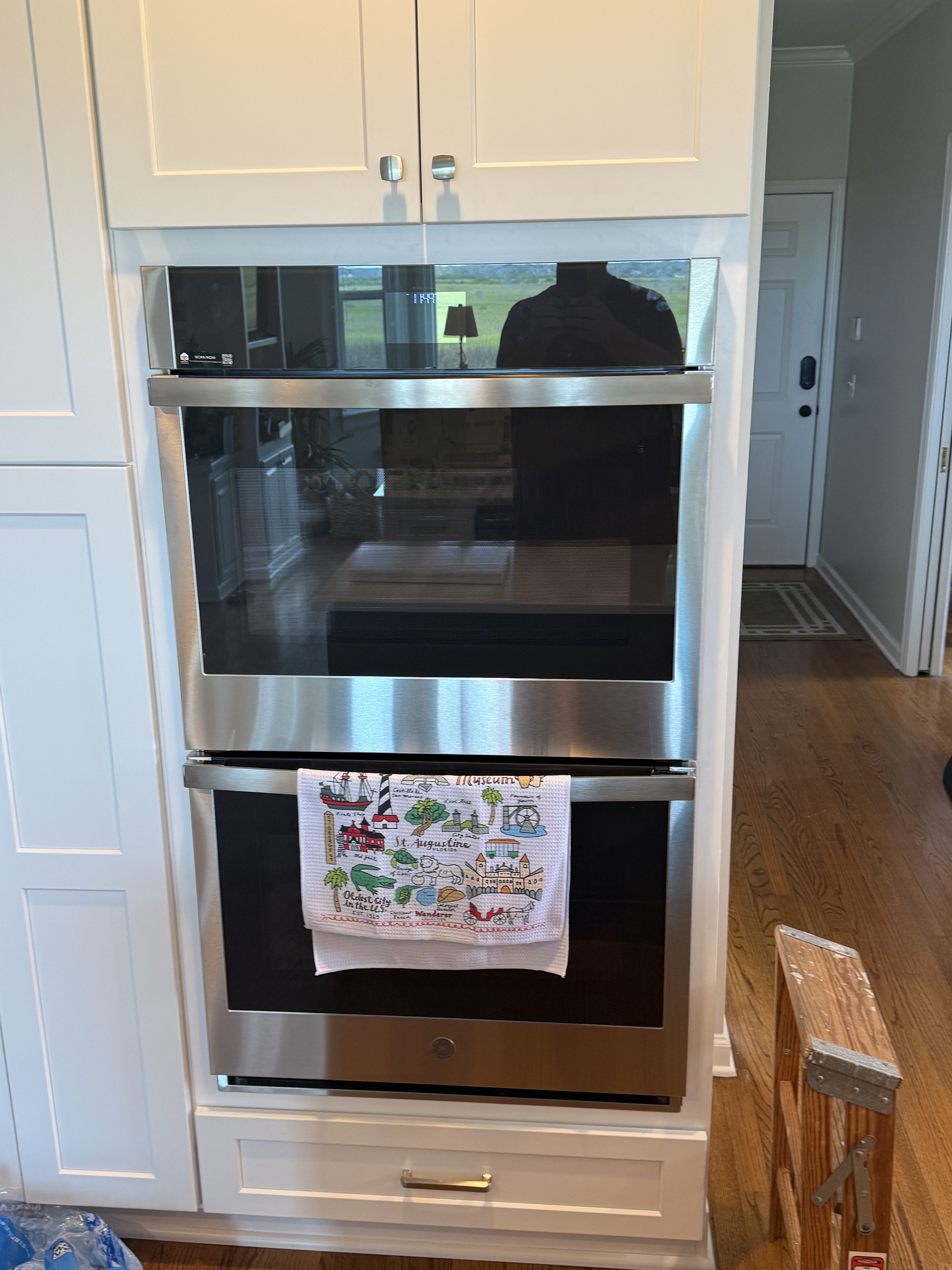 Stainless steel oven with a colorful towel hanging on the handle, positioned in a white kitchen with a wooden floor and a wooden step stool nearby.