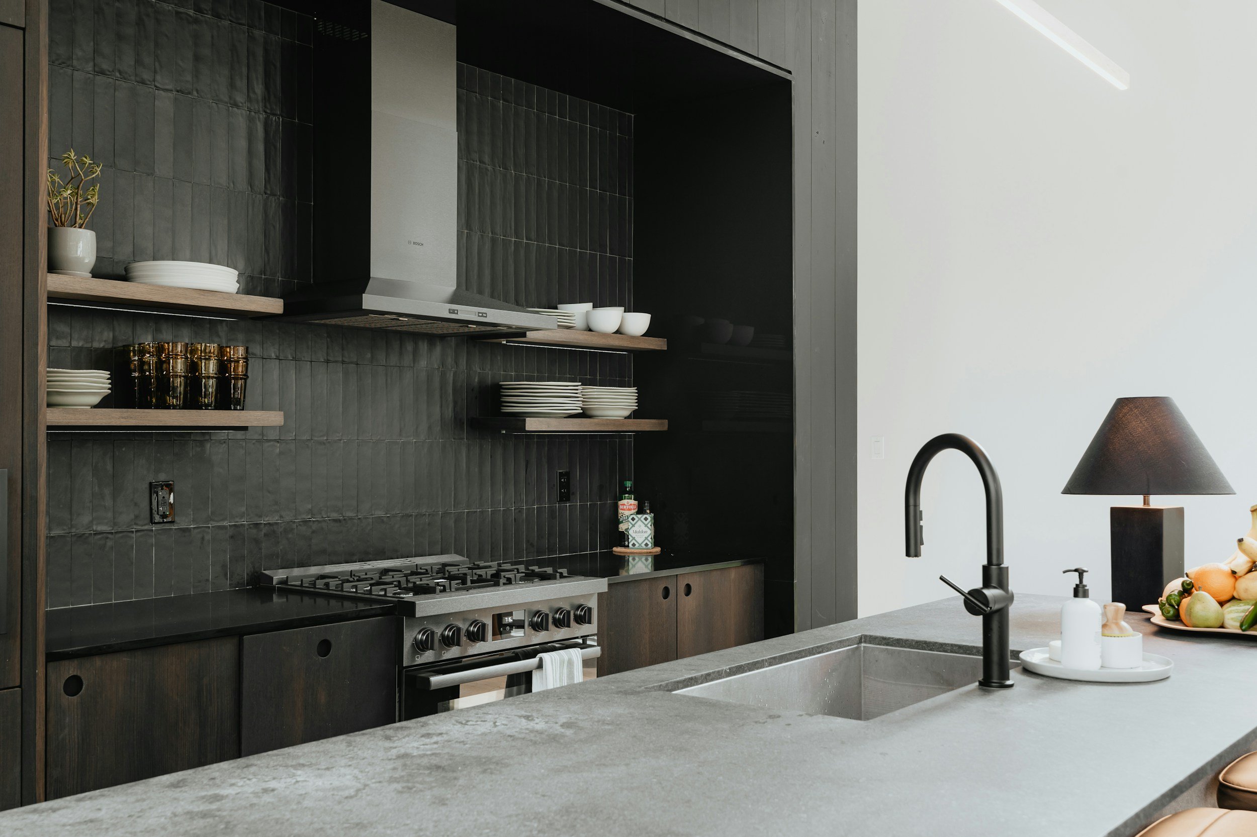 Modern kitchen with black tiled wall, built-in stove, open shelves with white dishes and glasses, and a gray countertop with a black faucet. There are fruits and a lamp on the counter.