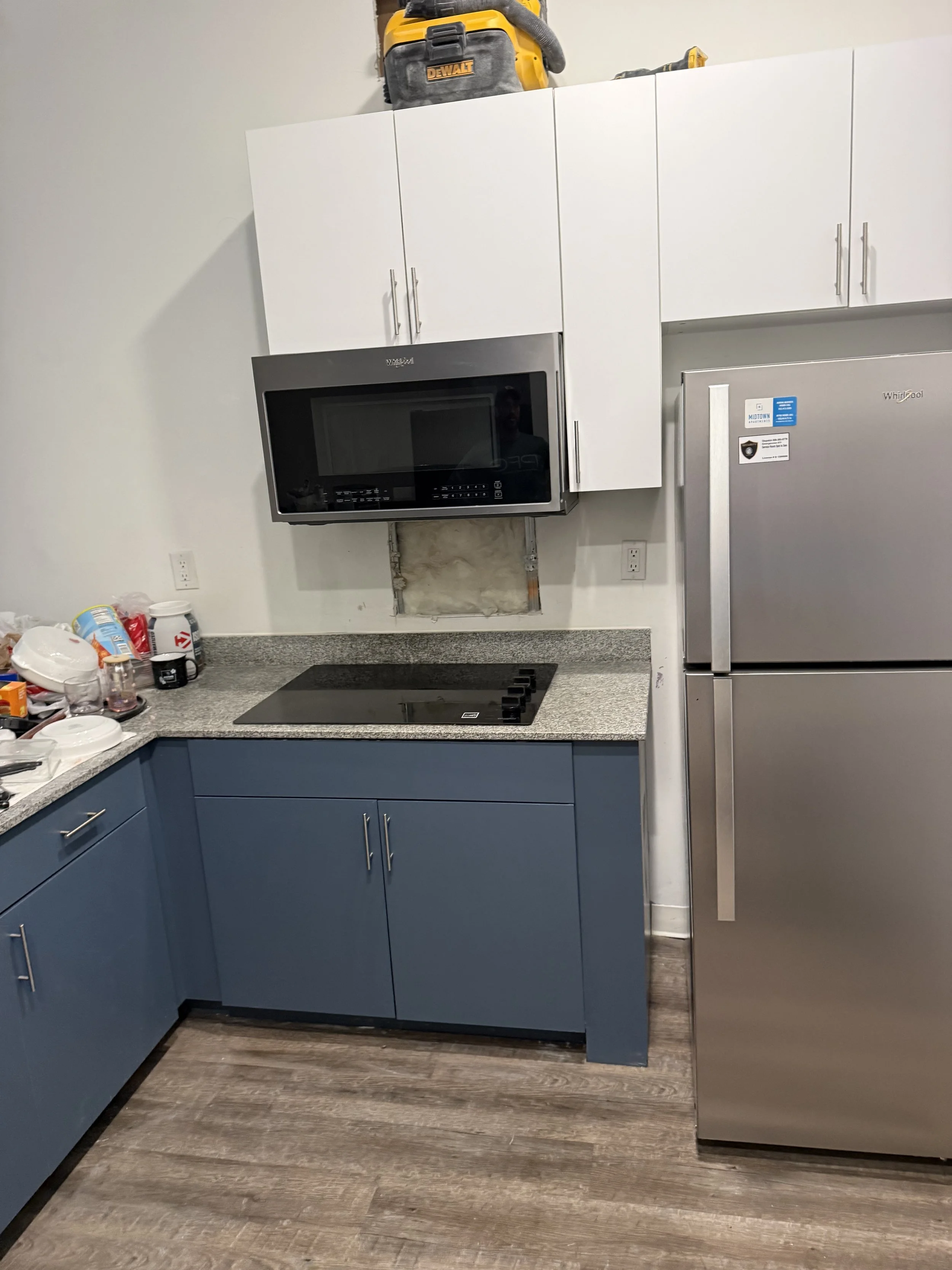 Kitchen with white upper cabinets, blue lower cabinets, gray countertop, microwave oven, stainless steel refrigerator, and miscellaneous items on the counter.
