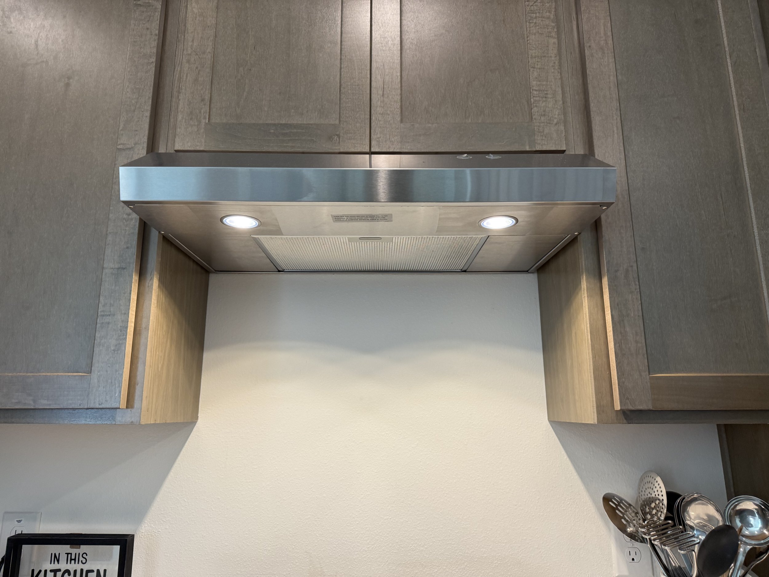 A kitchen with gray wood cabinets and a gray wall, with a stainless steel range hood and a small sign that reads 'In this kitchen'.