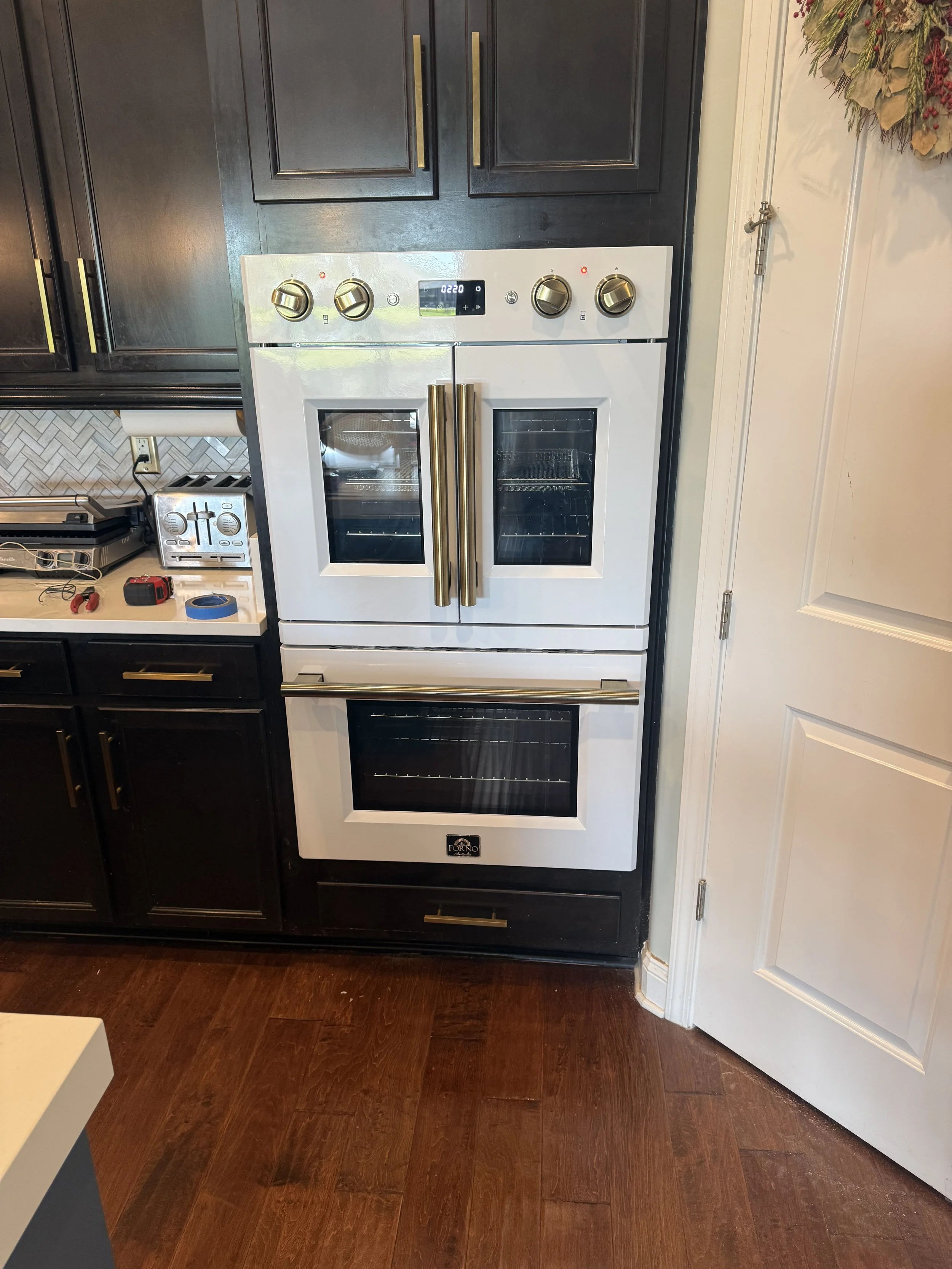 White oven with stainless steel handles, digital display, and four control knobs, situated between black cabinets with gold handles in a kitchen.