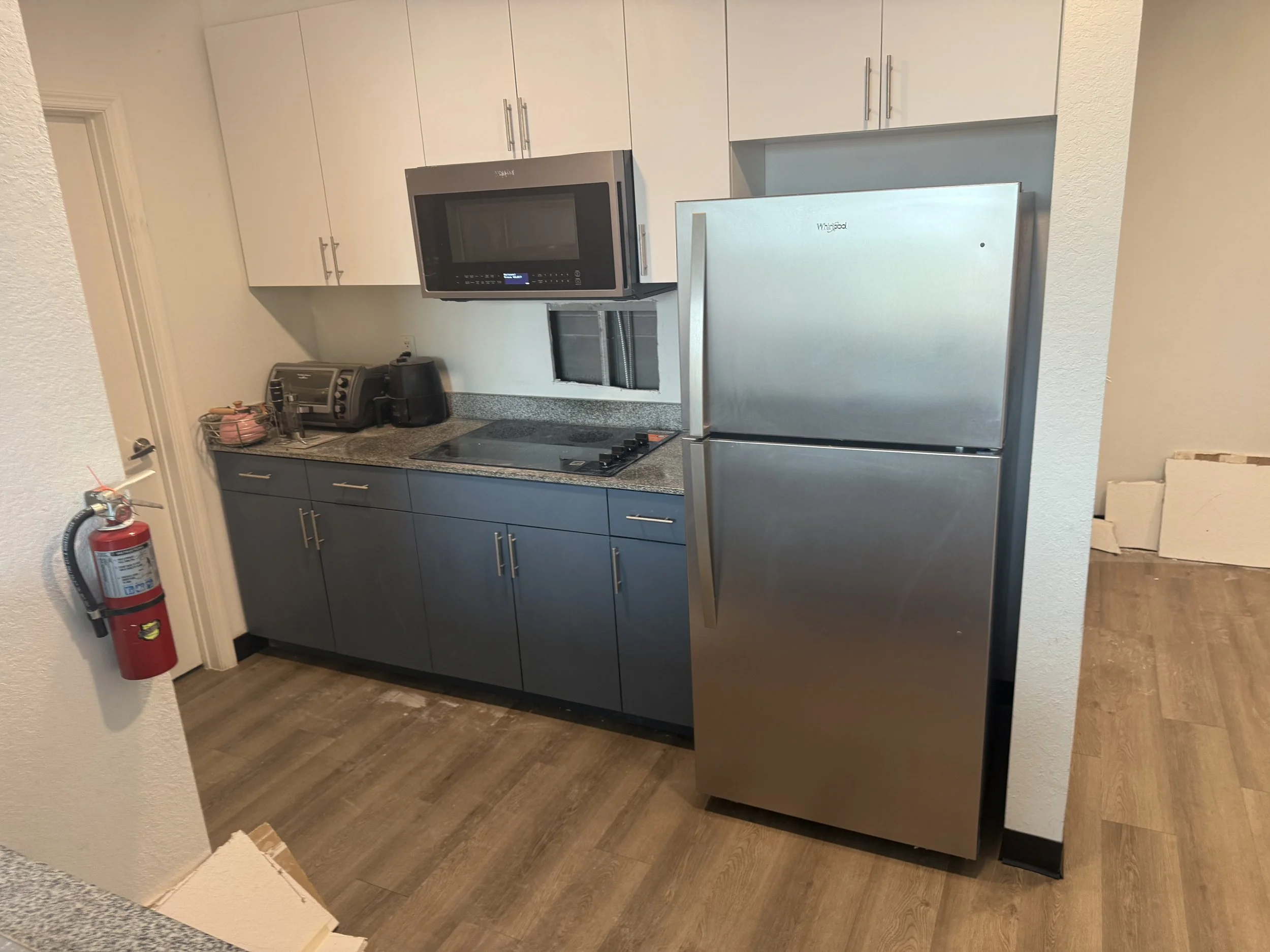 A kitchen with dark gray lower cabinets and white upper cabinets, a stainless steel refrigerator, an over-the-range microwave, a countertop with a toaster, a coffee maker, and other small appliances. There is a fire extinguisher mounted on the wall n
