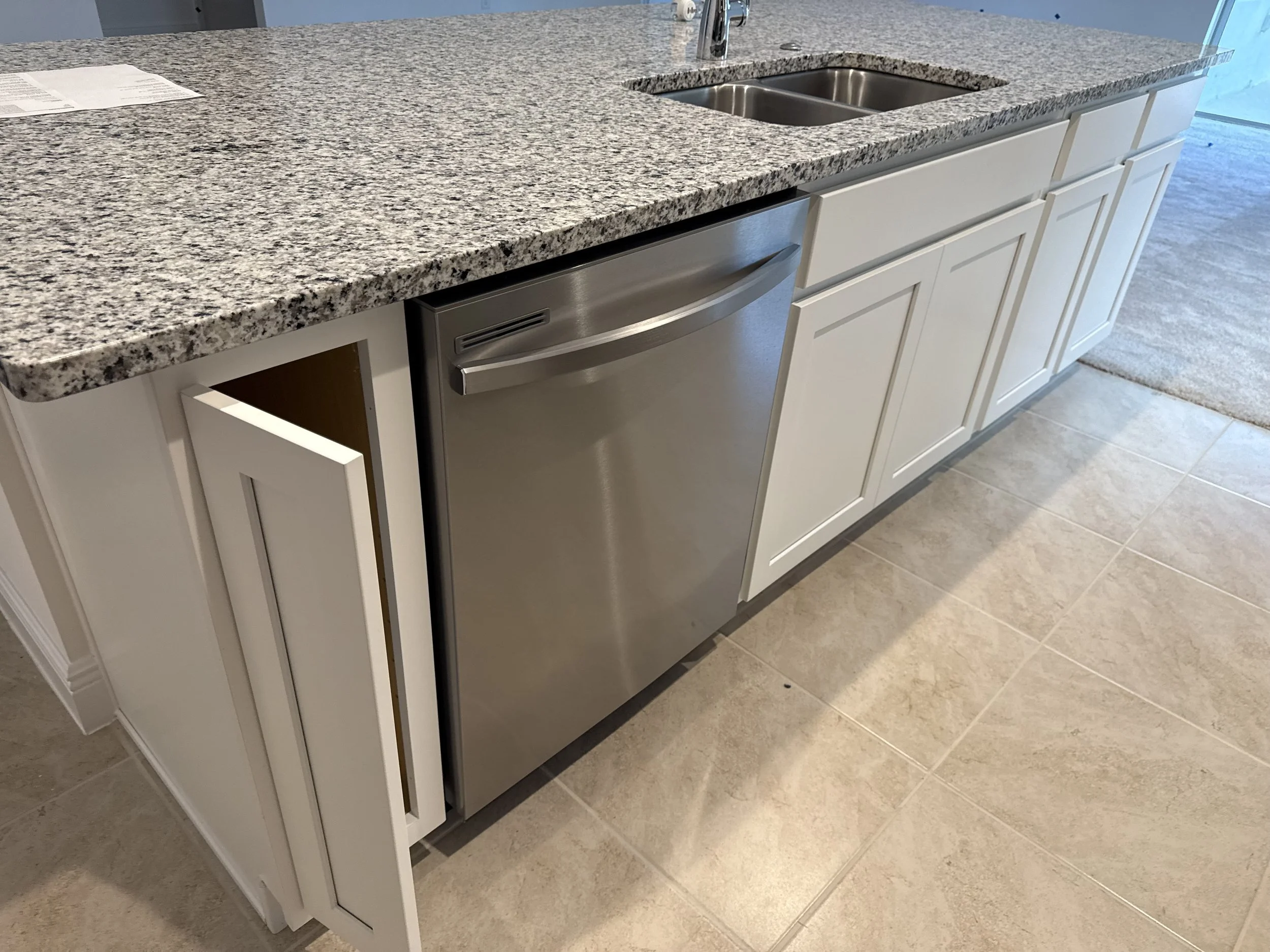 A kitchen countertop with a speckled granite surface, a double sink with a chrome faucet, white cabinets, a stainless steel dishwasher, a piece of paper, and a beige tiled floor.