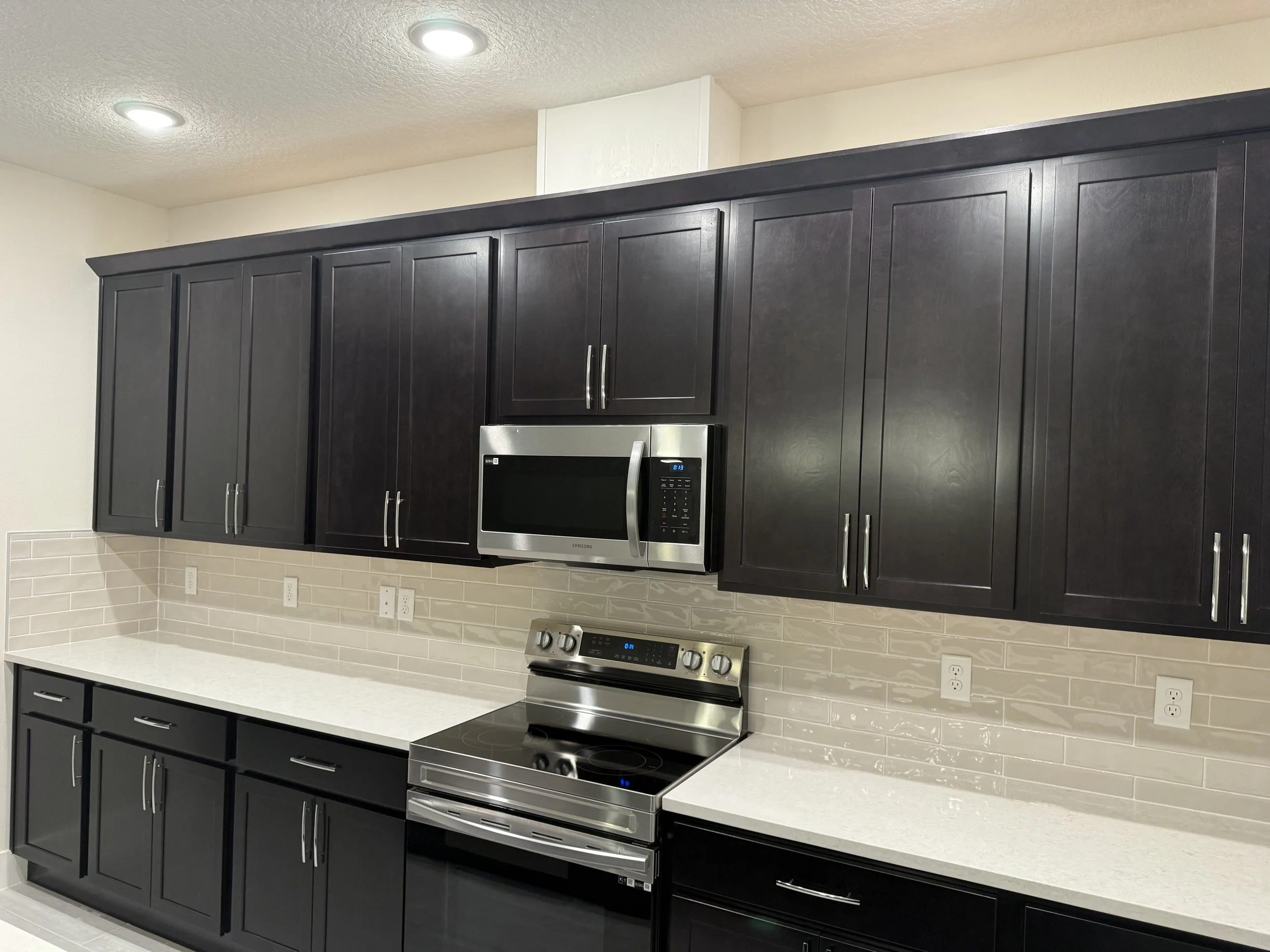 A modern kitchen with dark cabinets, a white countertop, a stainless steel microwave oven, and a stainless steel stove with an electric cooktop. There is a beige backsplash with subway tiles and multiple electrical outlets along the wall. The ceiling