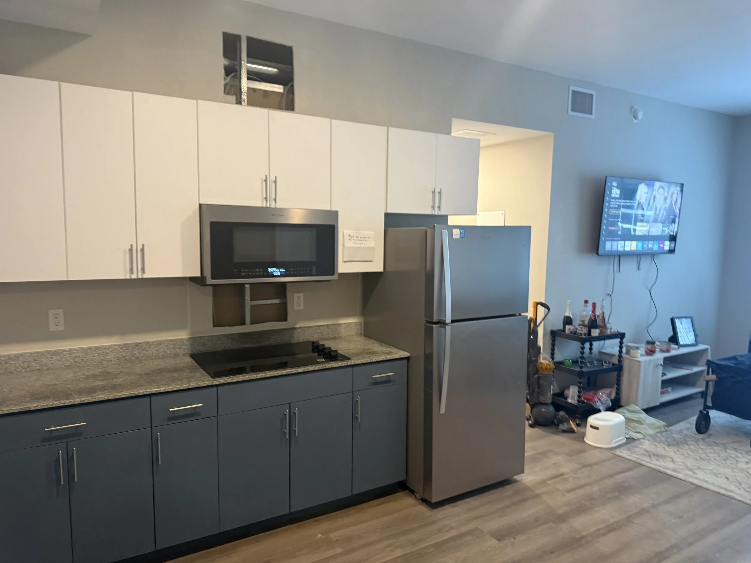 A kitchen with white upper cabinets, blue lower cabinets, a microwave oven, a refrigerator, a stove, and various small appliances and items on the counters and shelves.