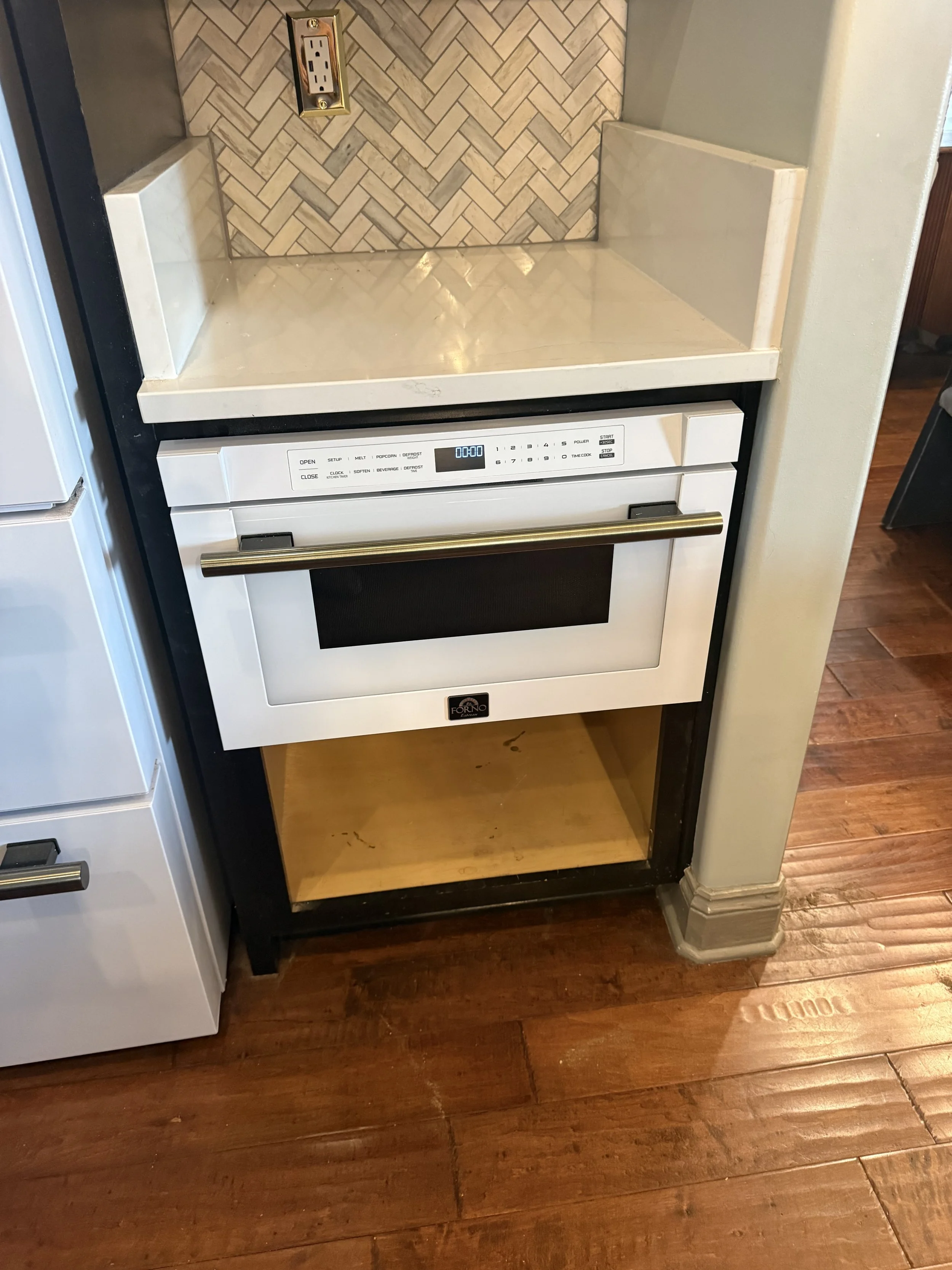 Kitchen area with a built-in oven and empty cabinet space above. The oven has a digital display and a brass handle. The backsplash features a herringbone tile pattern and an electrical outlet. The floor is hardwood.