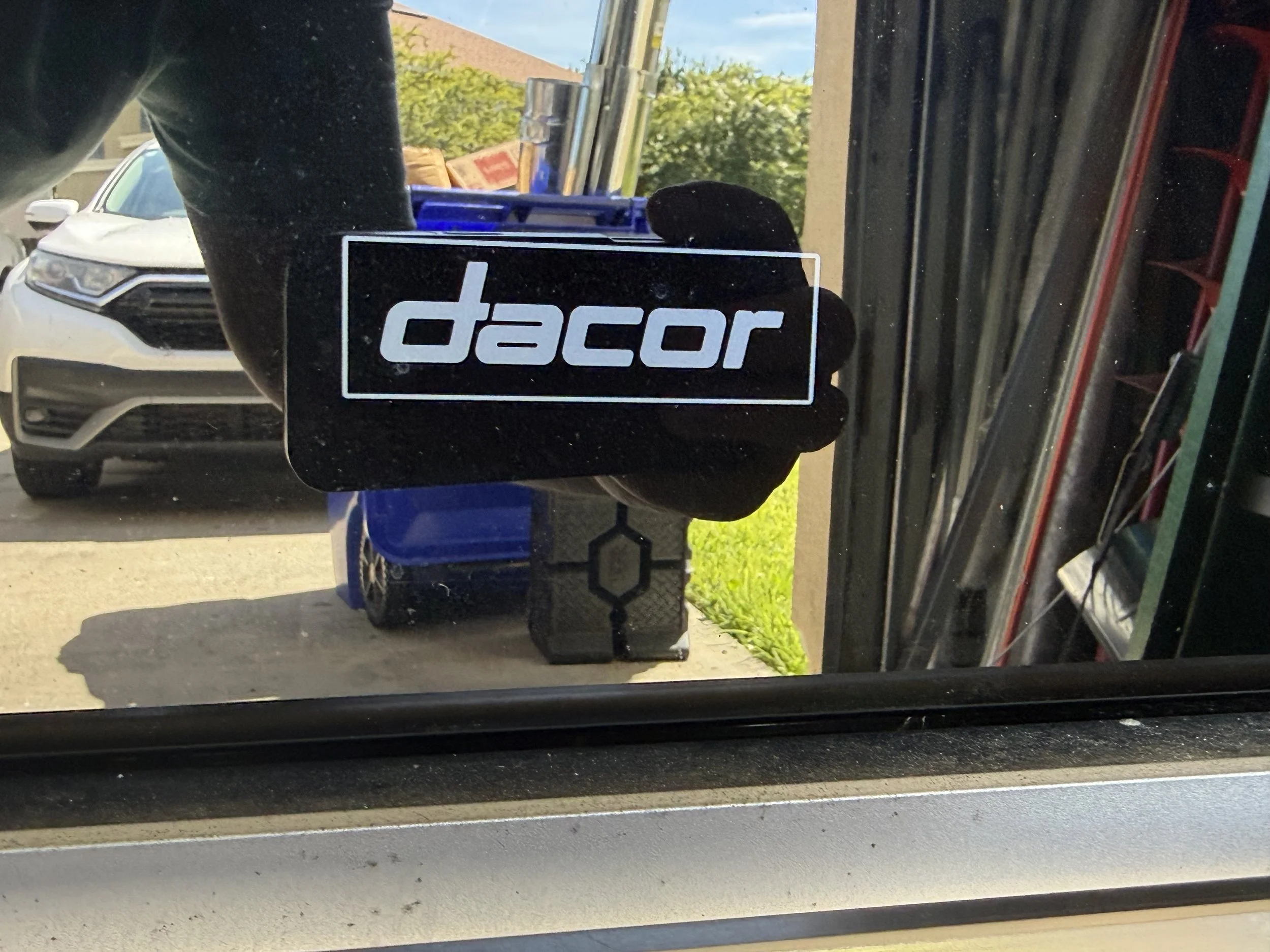 View through a car window showing a sticker that says 'dacar' on the reflection, with parked cars and greenery outside.