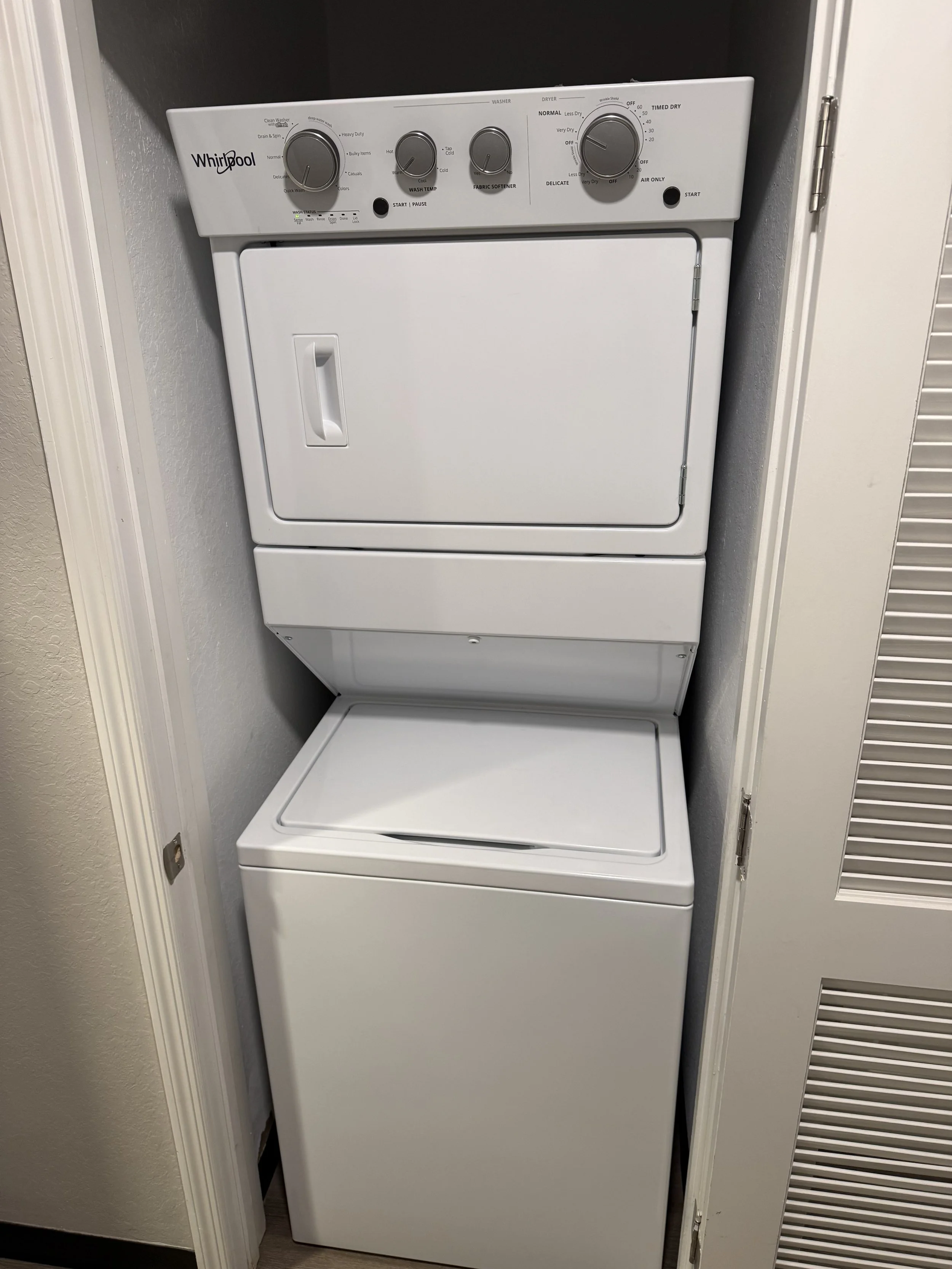 A stacked white clothes dryer on top of a matching white washing machine, located in a small laundry area with textured walls and a door frame to the left.