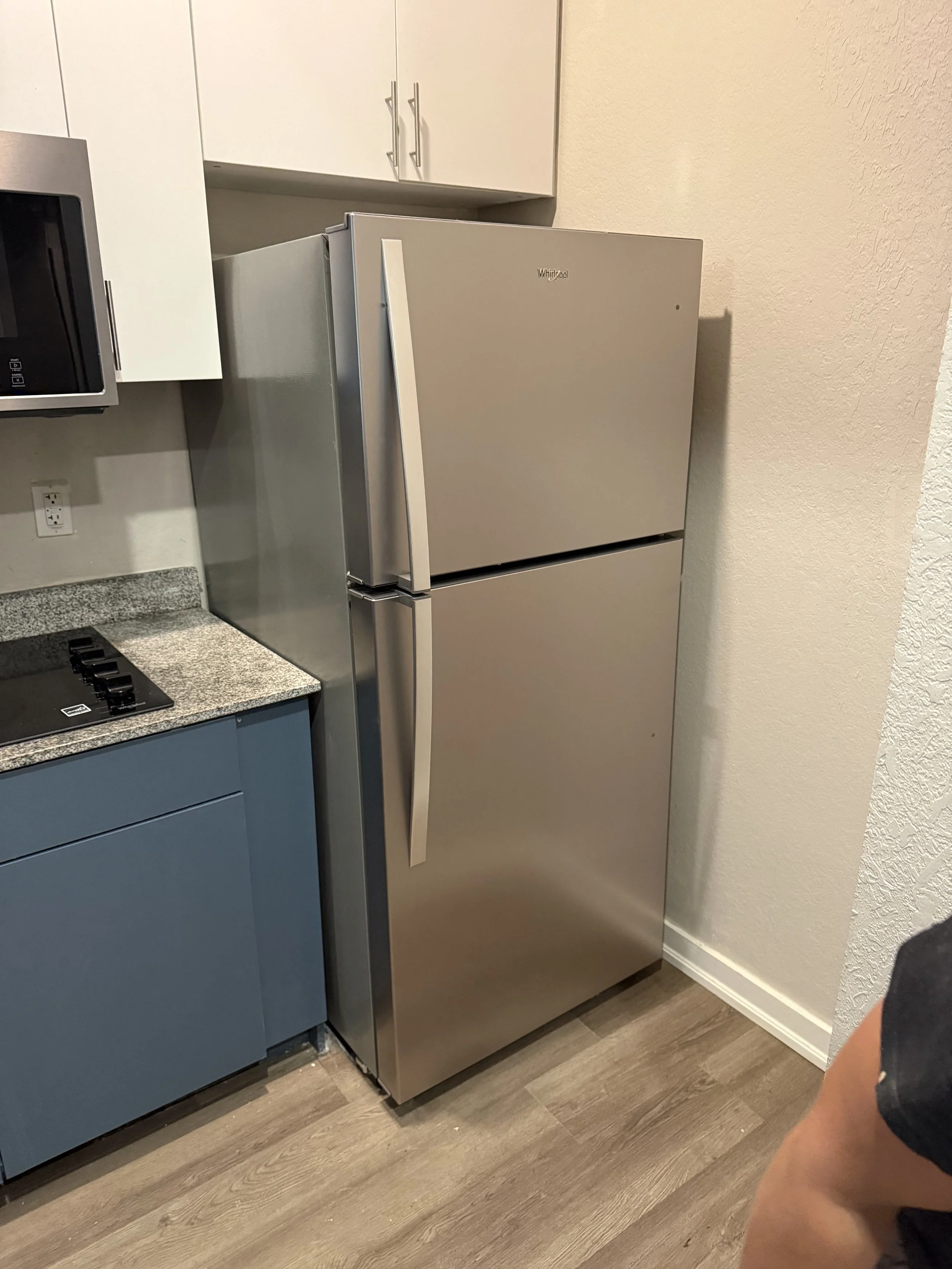 A modern kitchen corner with a stainless steel Whirlpool refrigerator next to blue kitchen cabinets and a black stove with a granite countertop. The floor is wood, and the wall has a textured beige finish.