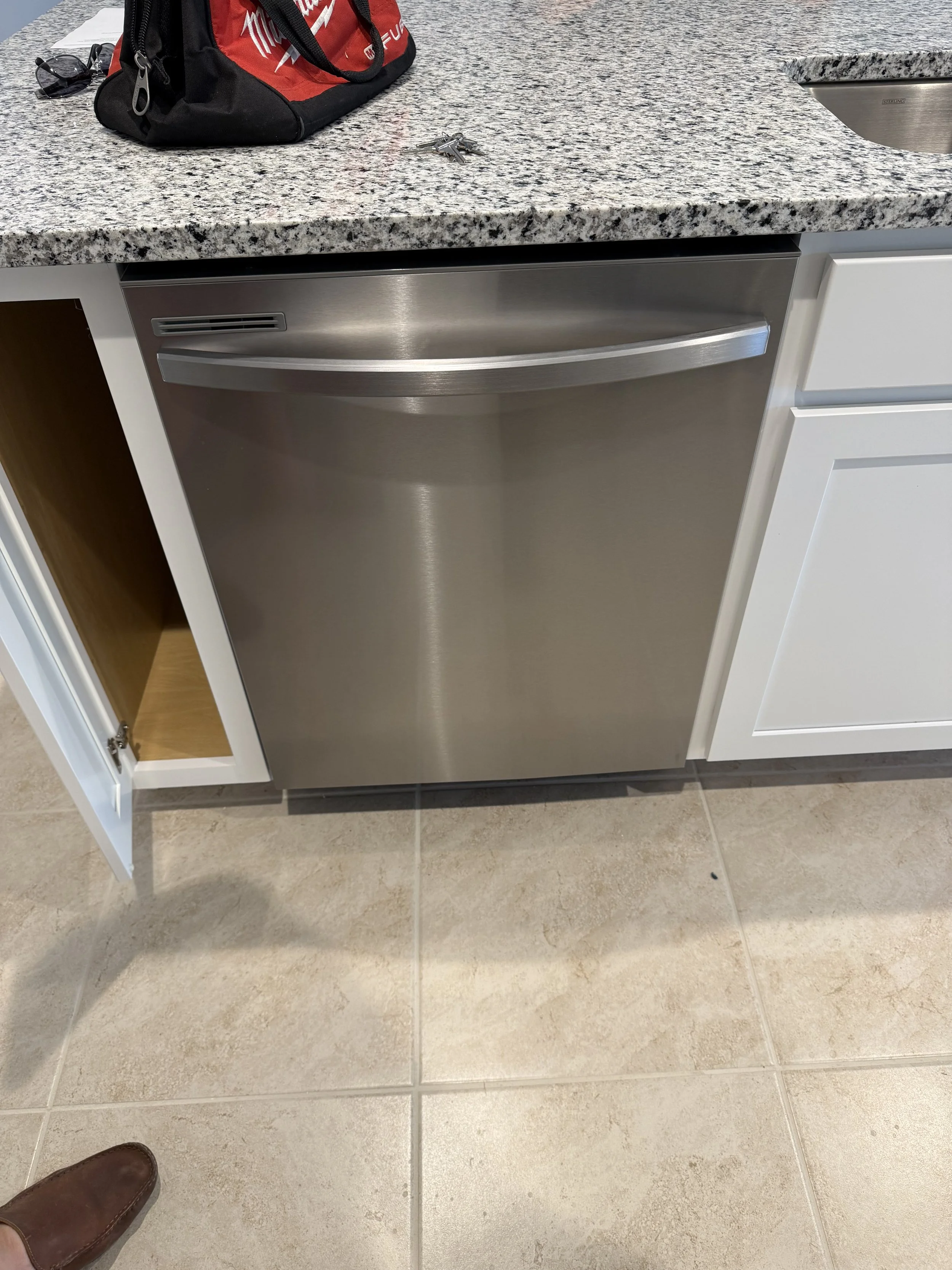 Kitchen counter with a red and black bag, a set of keys, sunglasses, and a paper. Below the counter is a stainless steel dishwasher and white cabinets. The floor is beige tile, and a brown shoe is visible at the bottom left corner.