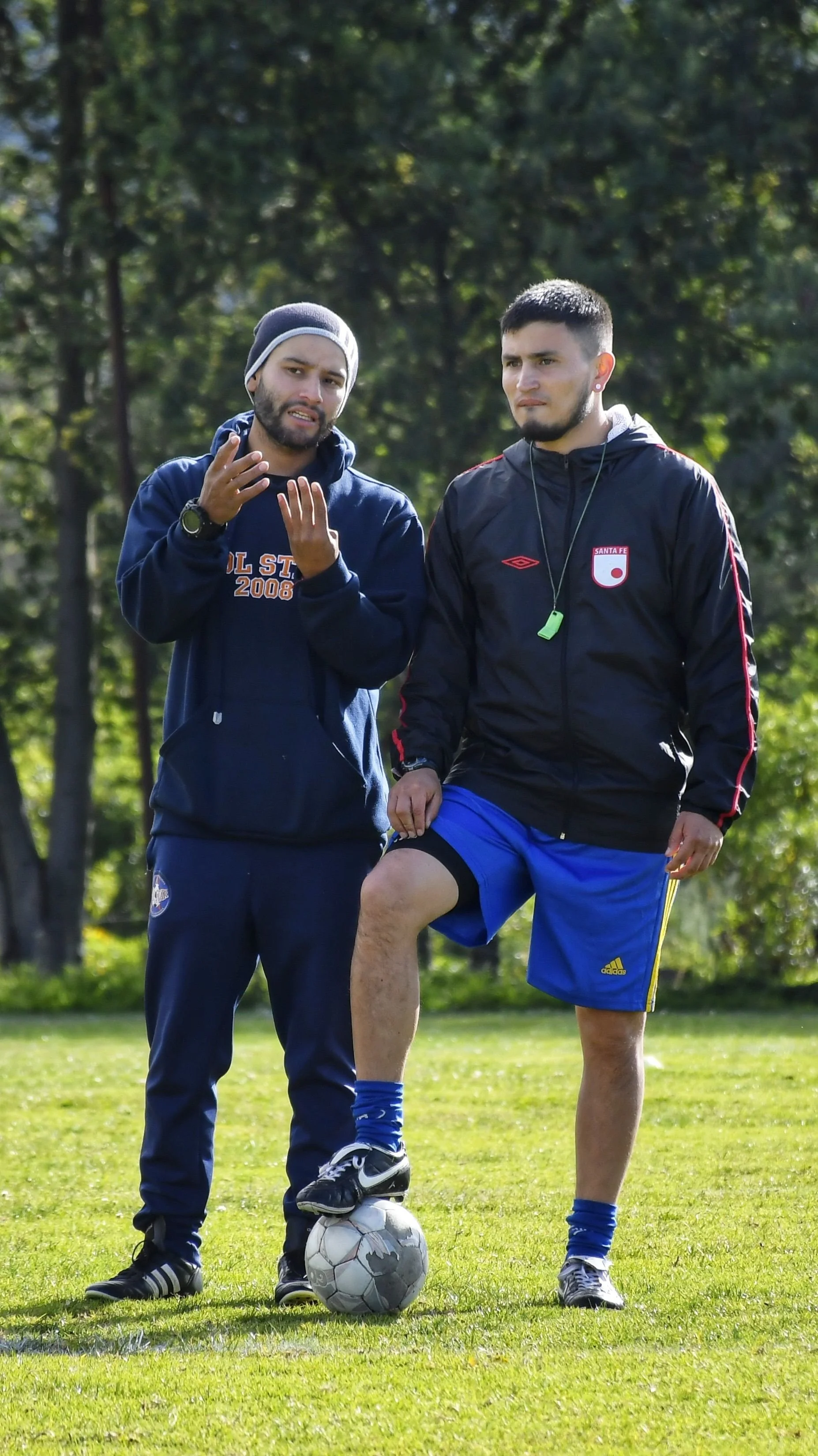 Two young men standing outdoors on a grassy field, one with a soccer ball and the other with one leg raised, engaging in a conversation or coaching session, with trees in the background.