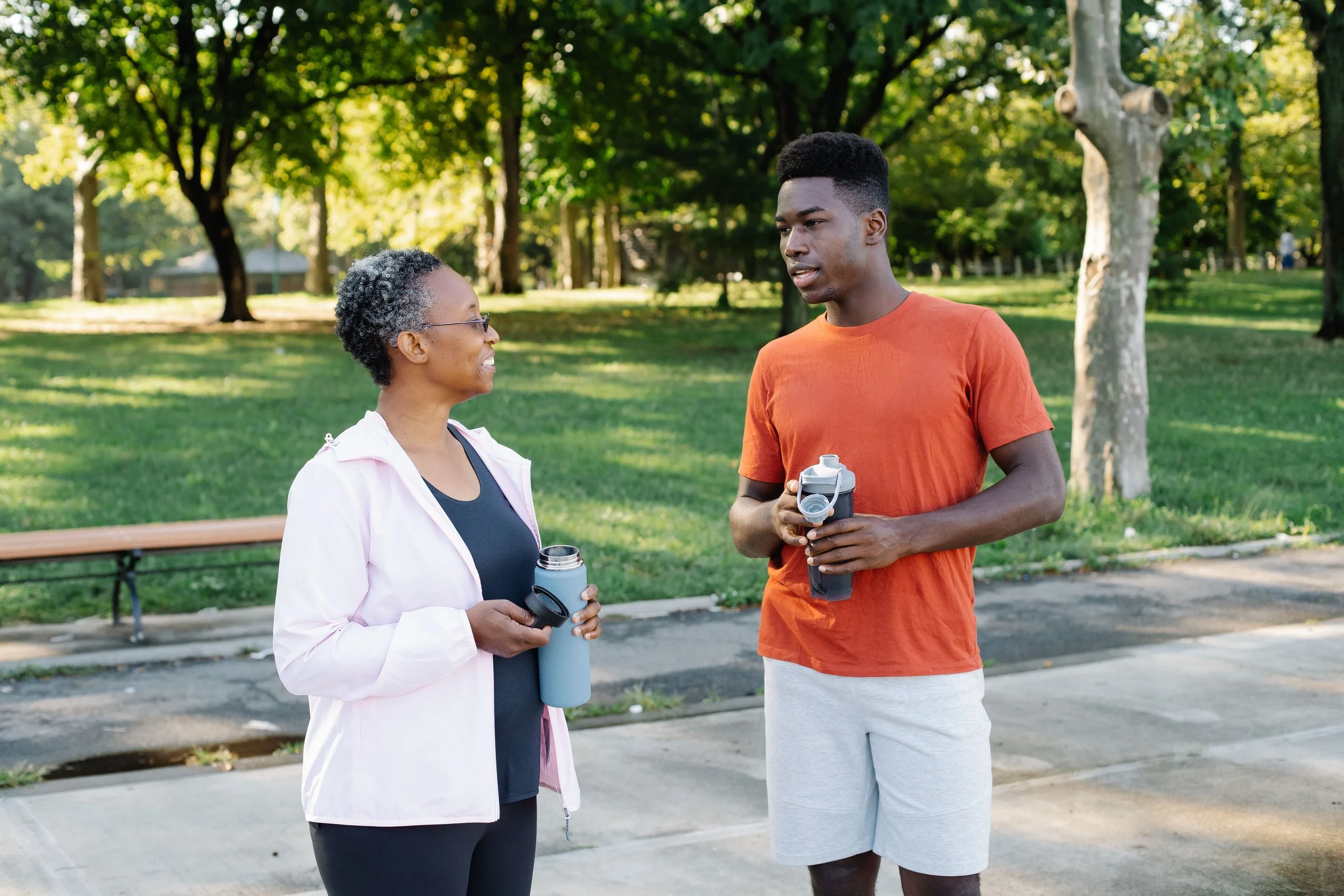 An older woman and a young man are talking in a park, both holding water bottles. The woman has short, gray curly hair and glasses, wearing a white jacket over a dark shirt. The young man has a tall, flat-top hairstyle, wearing an orange T-shirt and gray shorts. They are smiling and engaging in conversation, with trees and a bench visible in the background.