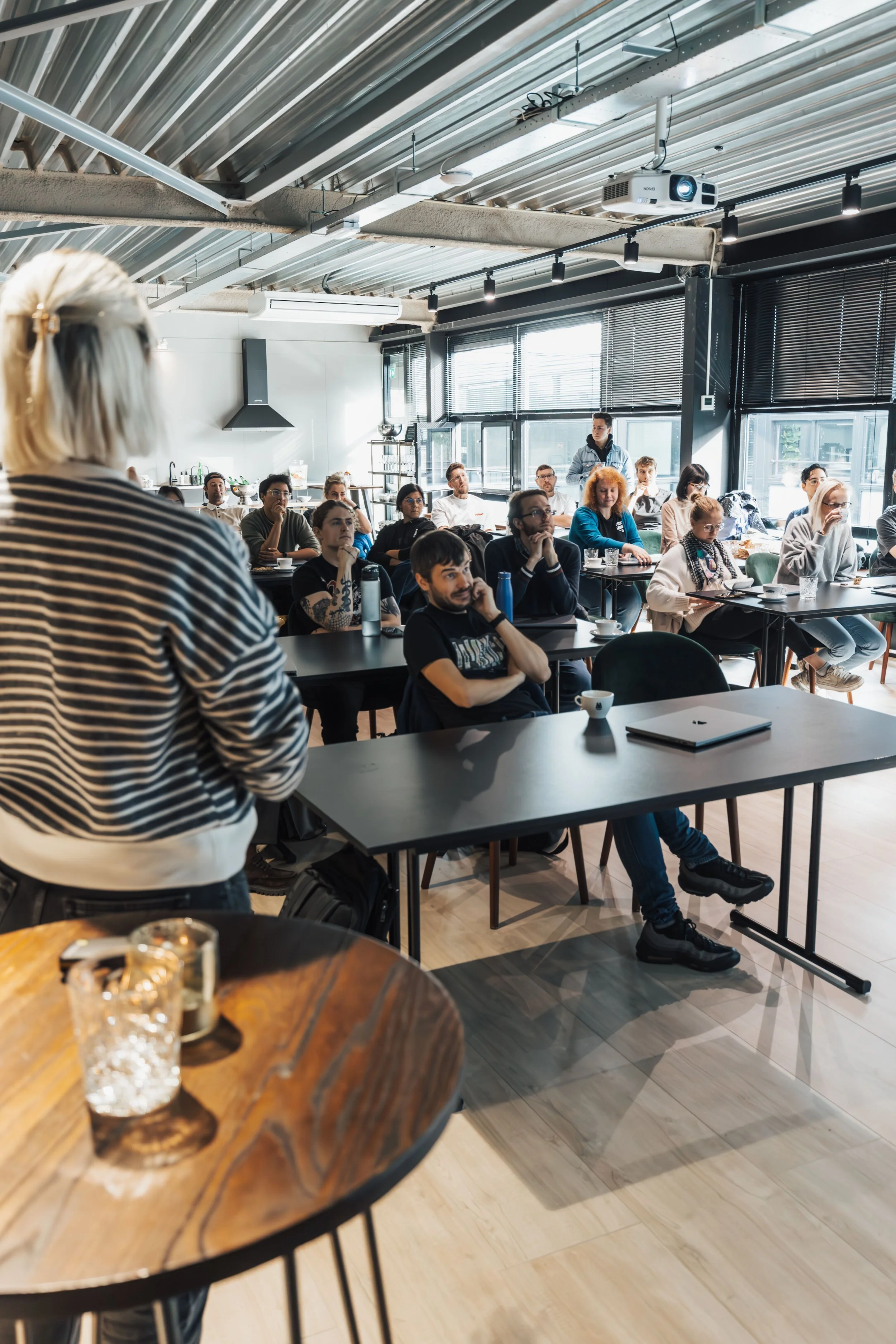 A group of people attending a lecture or presentation in a modern, industrial-style conference room with large windows, blinds, and exposed ceiling pipes. A woman with blonde hair in the foreground is speaking or presenting, while the audience is seated and listening attentively.