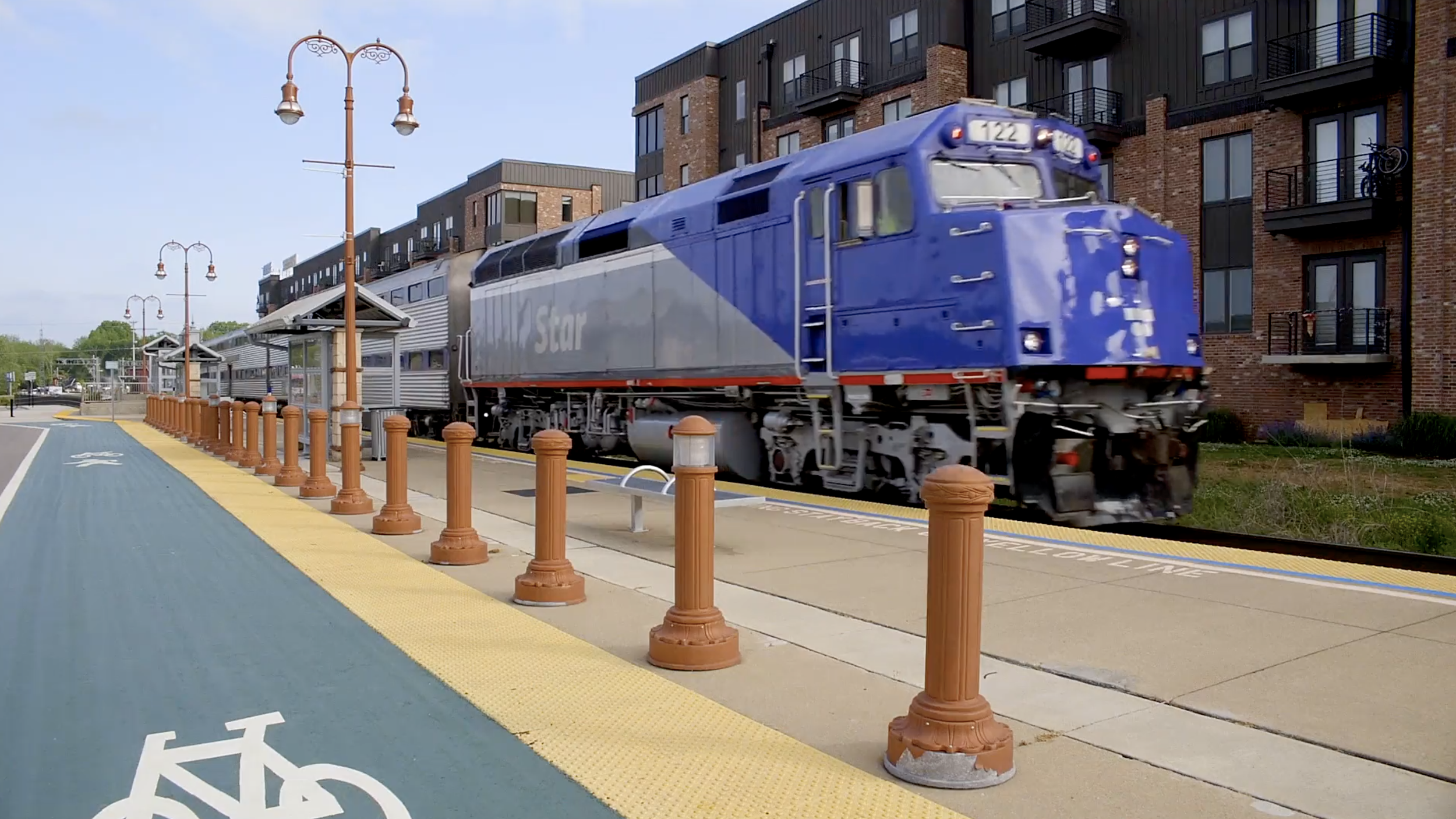 A blue and gray train with the words 'Amtrak Star' on its side, moving along a street with modern apartment buildings in the background. There are orange safety posts along the sidewalk and street lamps overhead.