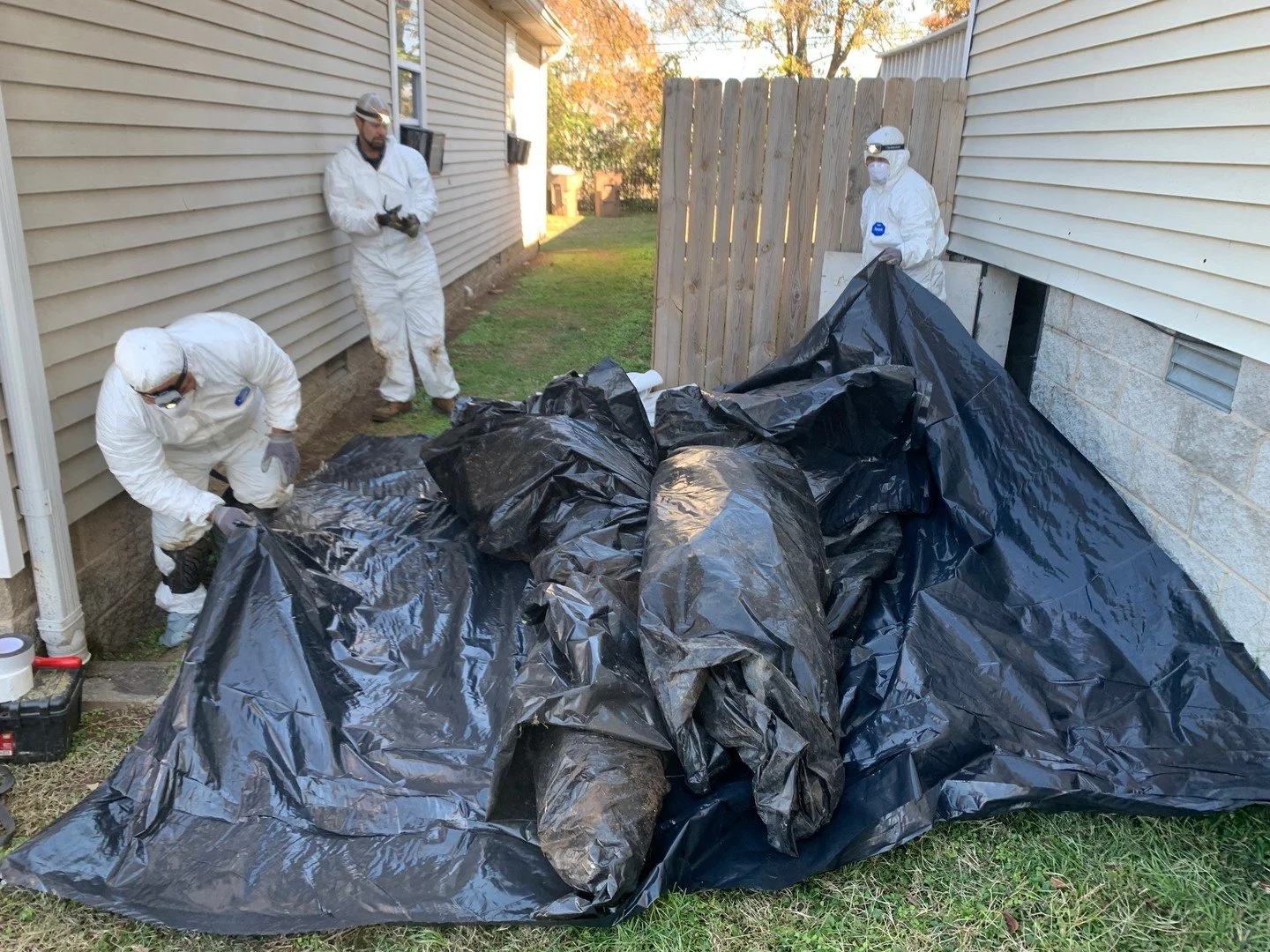 The old material has to physically leave the property. That's a loaded tarp bag coming out of the crawl space access in the foundation, dragged through the gap between the siding and the fence by three people. Hauling is the part of Insulation Restor