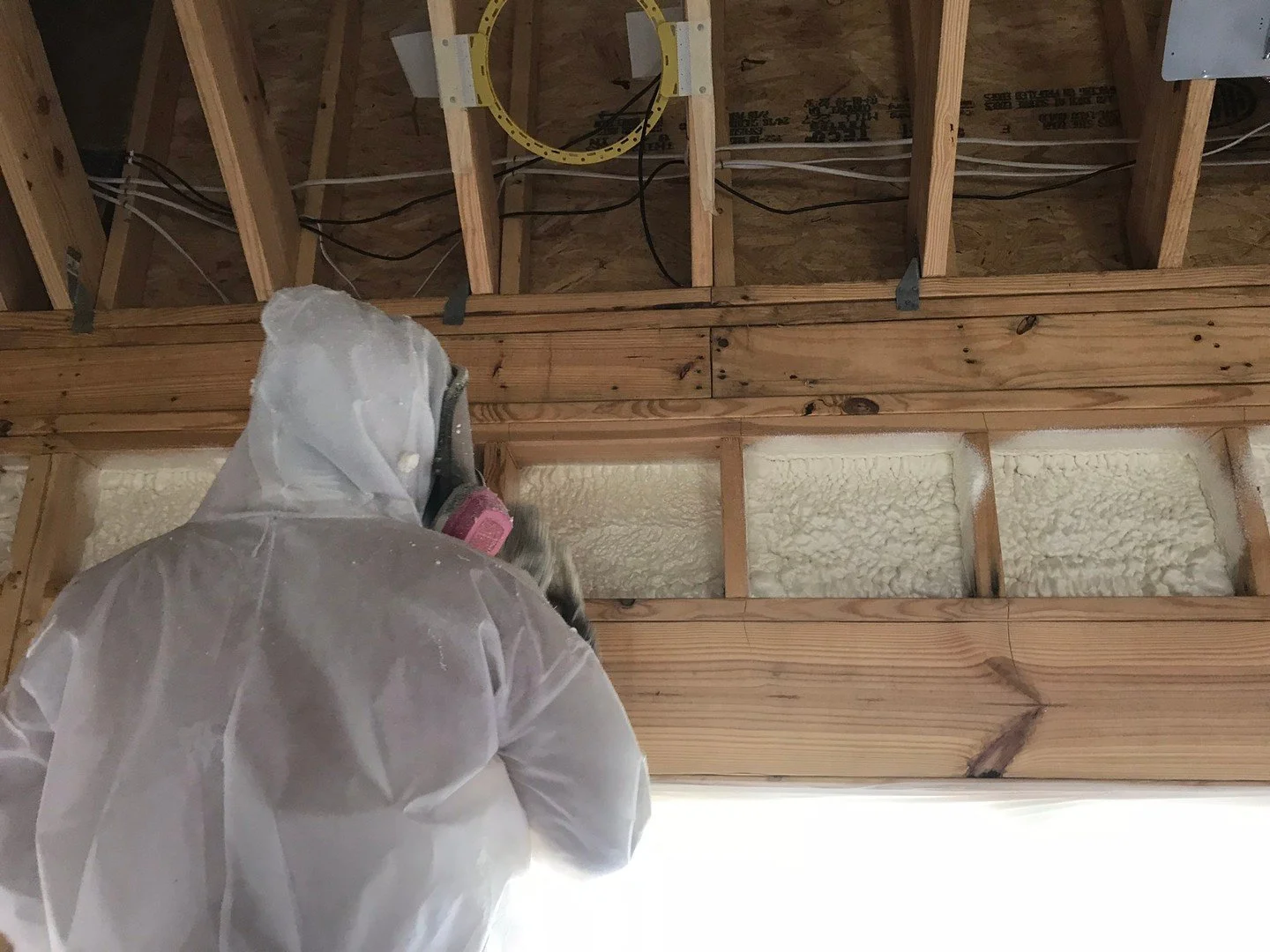 Closed cell foam under a roof deck, new construction, sprayed one rafter bay at a time. Two bays over are already full and set, the bay overhead is still bare wood, and the framing between them stays exposed. Closed cell goes on dense and holds tight
