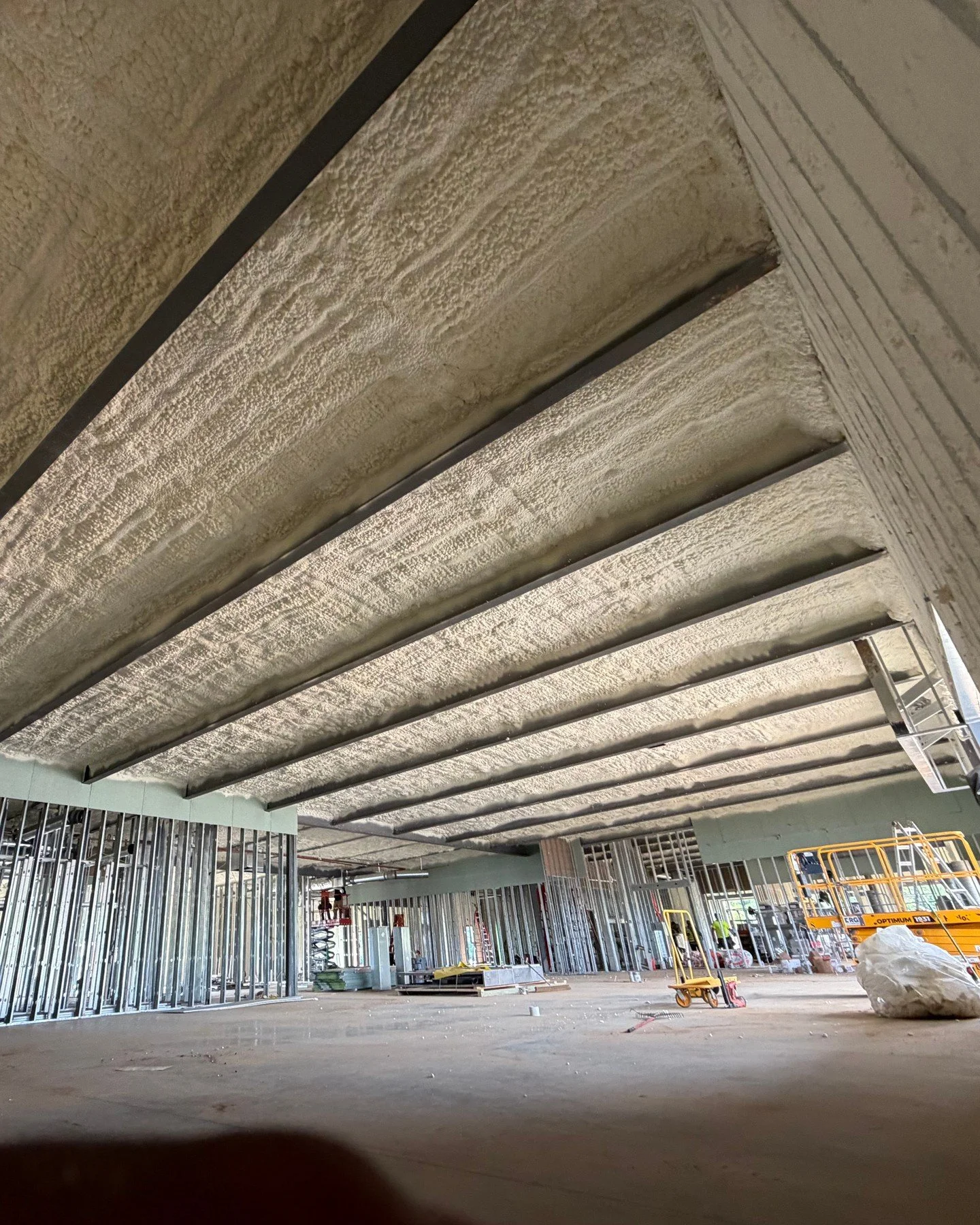 Open cell on the roof deck of a truck stop under construction, every purlin bay sprayed eave to ridge with no breaks. On commercial work the builder writes the spec and the install gets done to it, nothing improvised on site. The scissor lift is stil