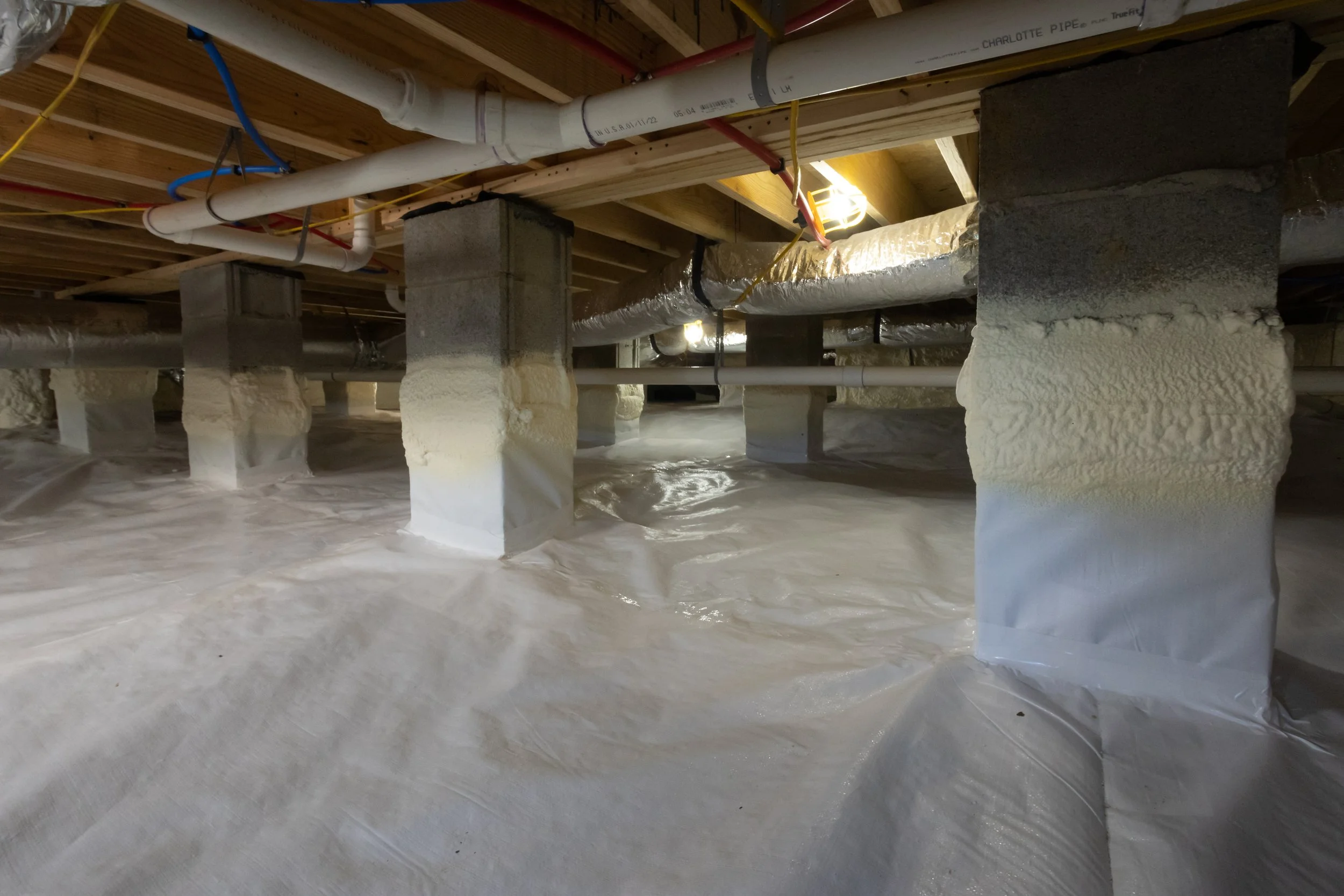 Insulation foam sprayed on crawl space, with wooden beam ceiling and plastic vapor barrier.