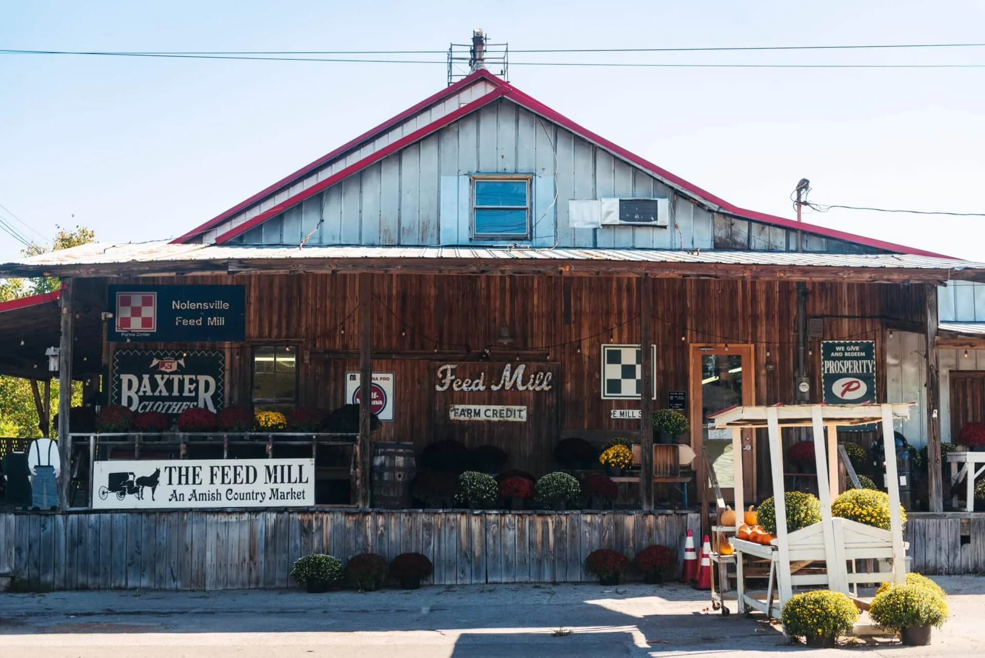 A rustic wooden building with a sign that reads "The Feed Mill," advertising an Amish country market. The building has a red roof and a small storefront decorated with potted plants and pumpkins outside.