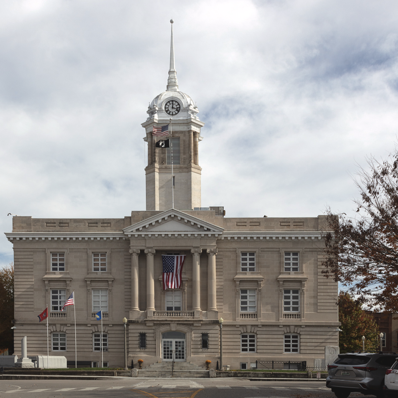 Front view of a government building with a clock tower and flags, including the American flag, on a partly cloudy day.