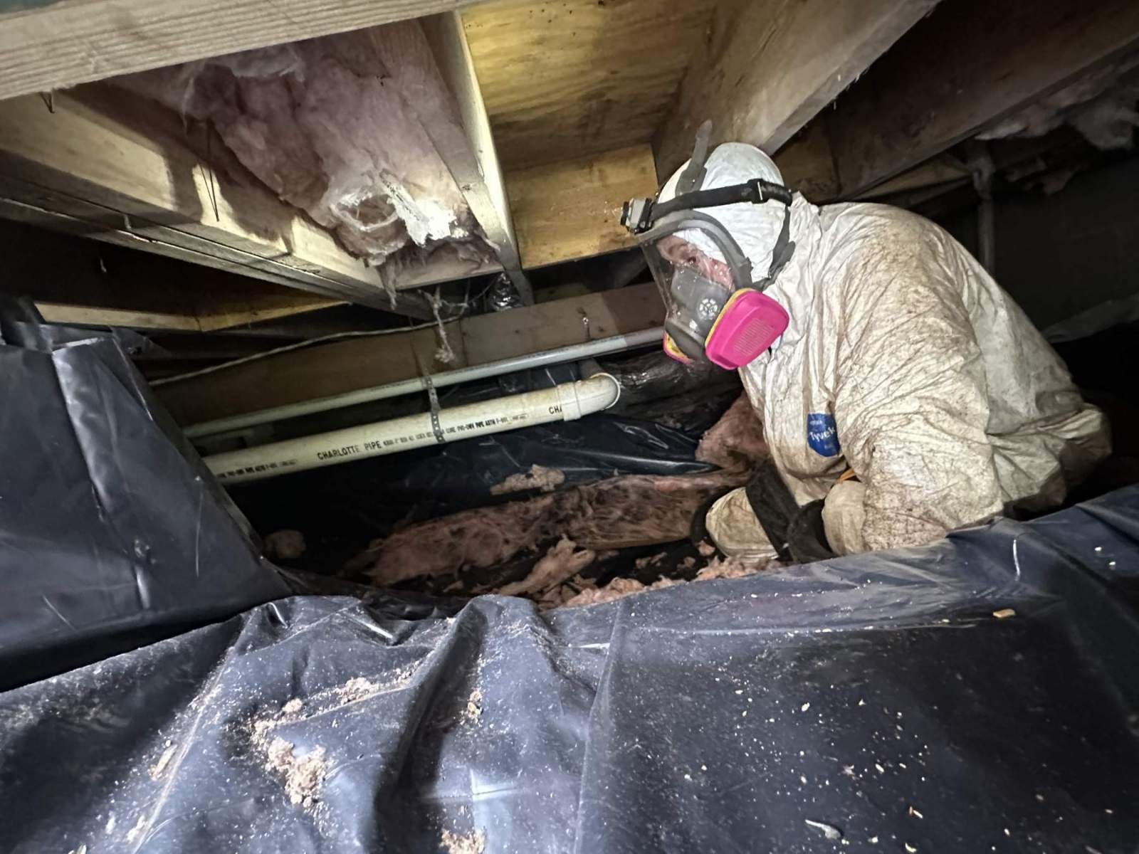 A person wearing protective gear and a face mask inspecting the space beneath a house or attic with exposed insulation and plumbing.